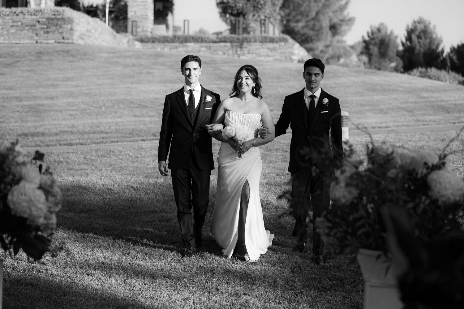 Black and white wedding photo showing three people walking together on a grassy hillside at an outdoor ceremony.