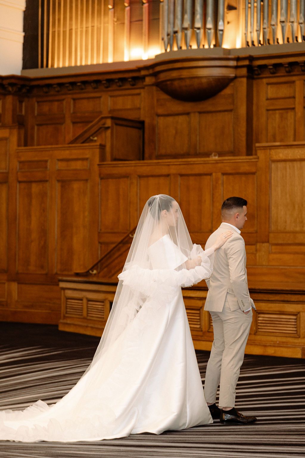 Wedding couple in white attire photographed against wooden paneled walls in warm lighting.