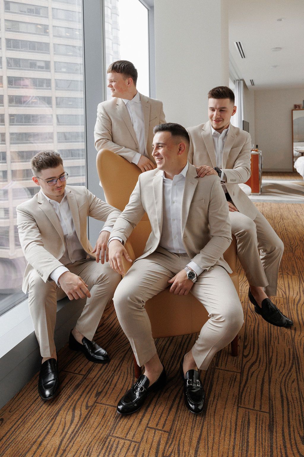 Groomsmen in light beige suits lounging on a brown couch in a modern room with city views.