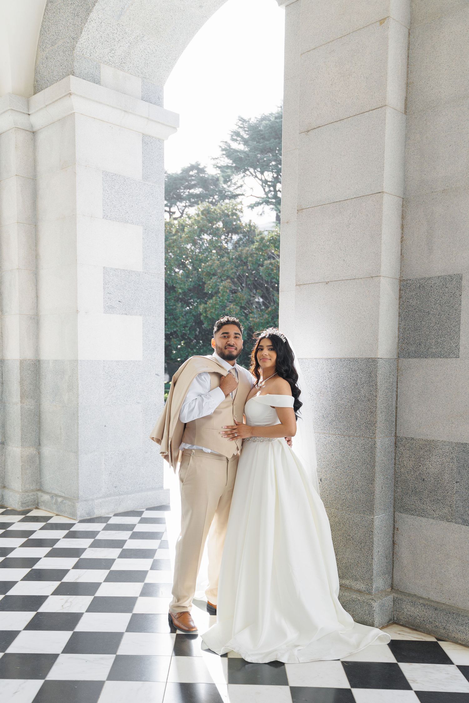 Wedding couple dances on black and white checkered floor under white columned archway.