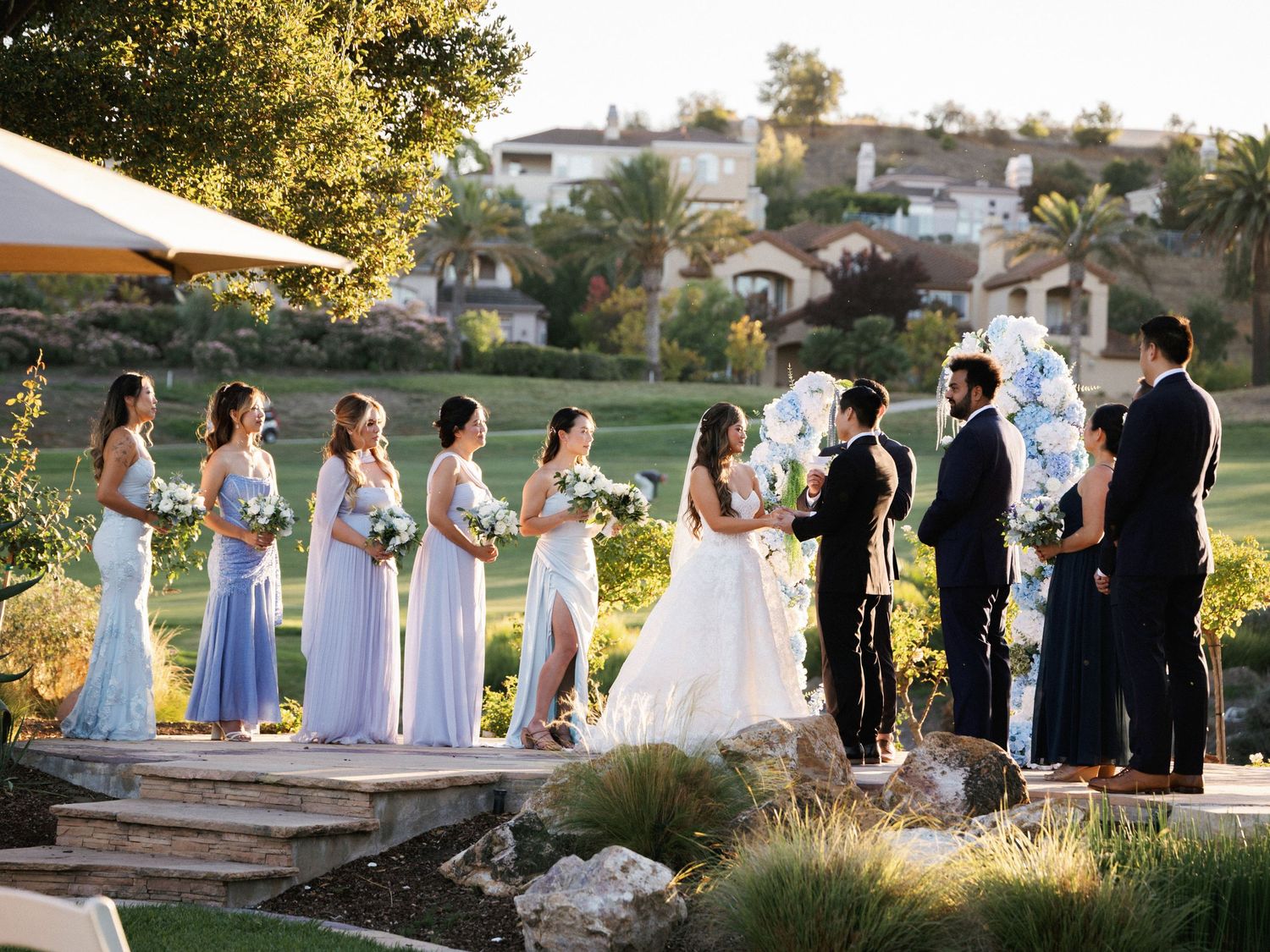 Wedding ceremony taking place outdoors with bridal party in blue dresses against a scenic hillside backdrop.