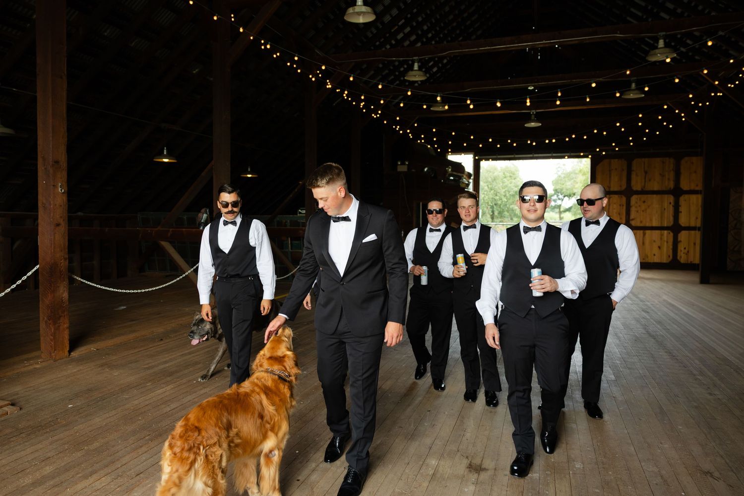 A wedding party walks across a rustic wooden floor decorated with string lights as a golden retriever leads the way.