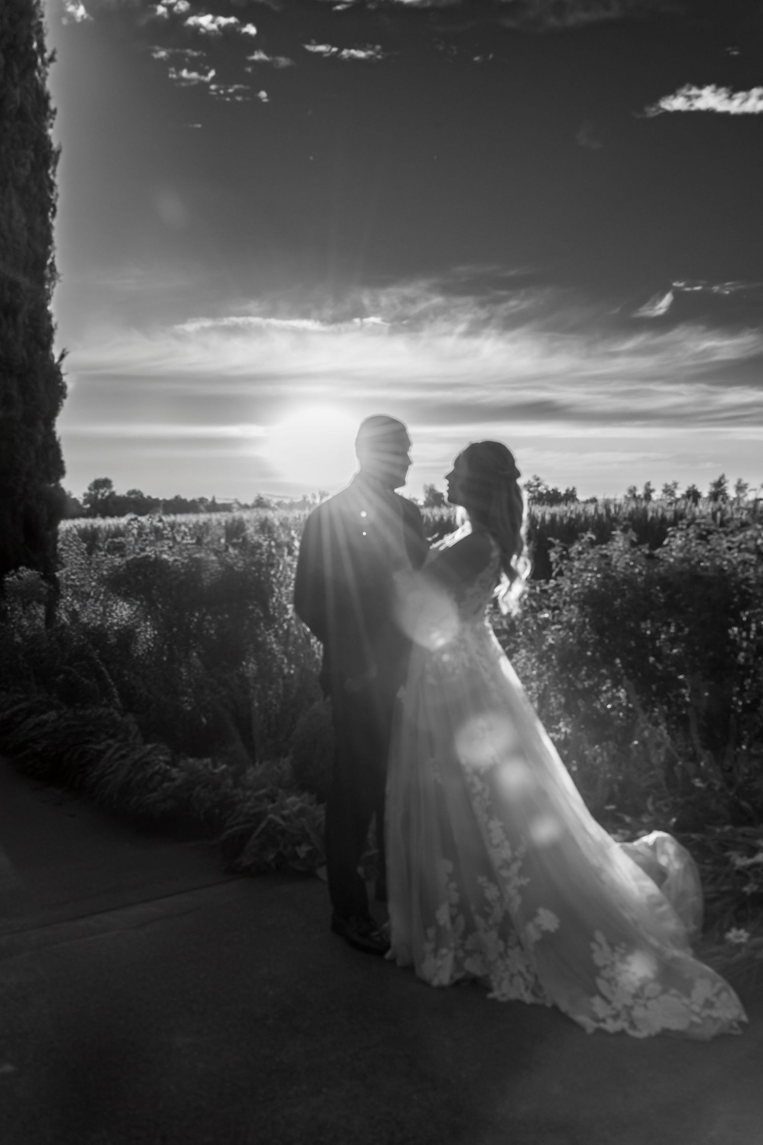 Silhouetted couple in wedding attire embrace against dramatic sunset sky with lens flare.
