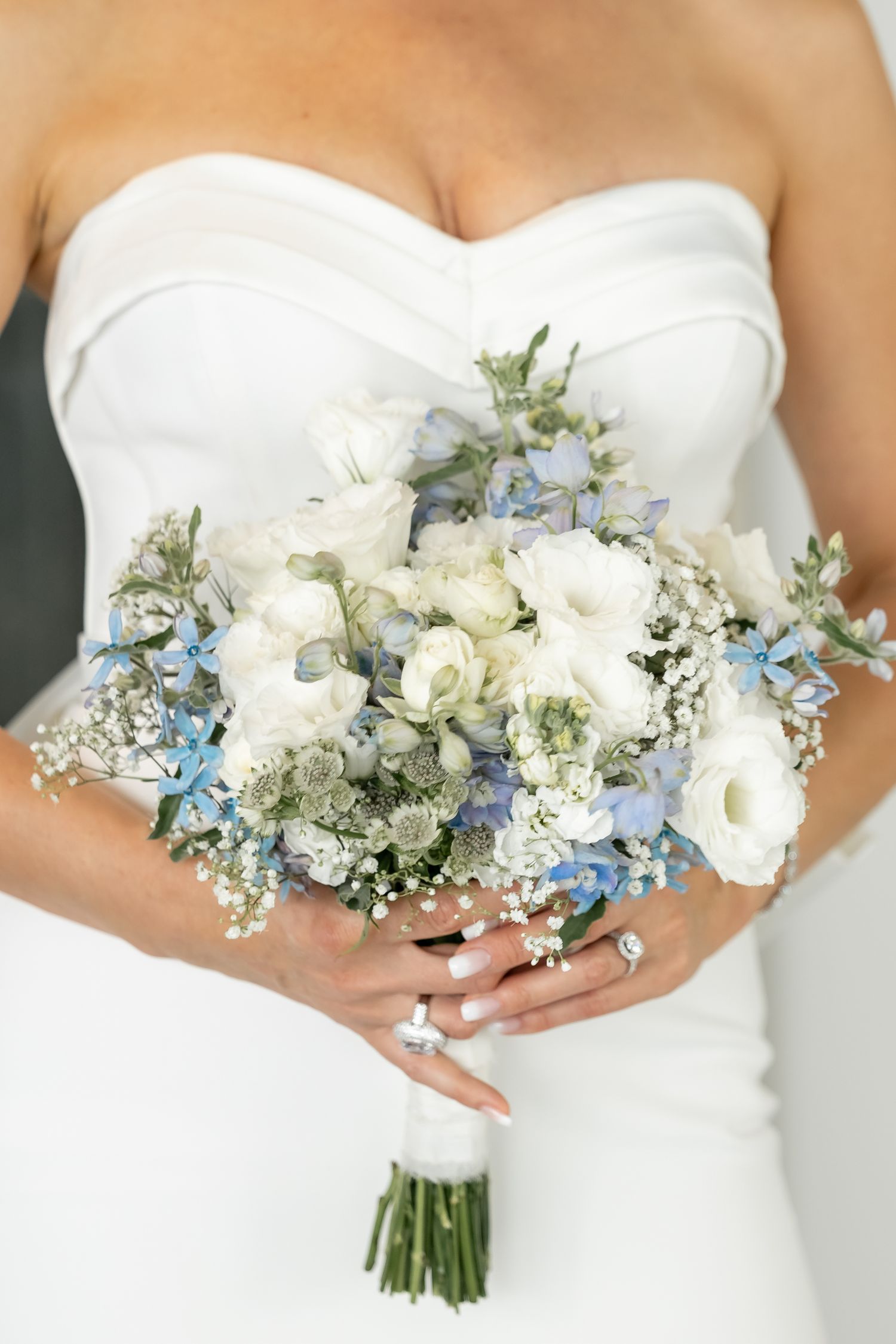 White bridal bouquet featuring calla lilies, baby's breath and delicate blue flowers against a light background.