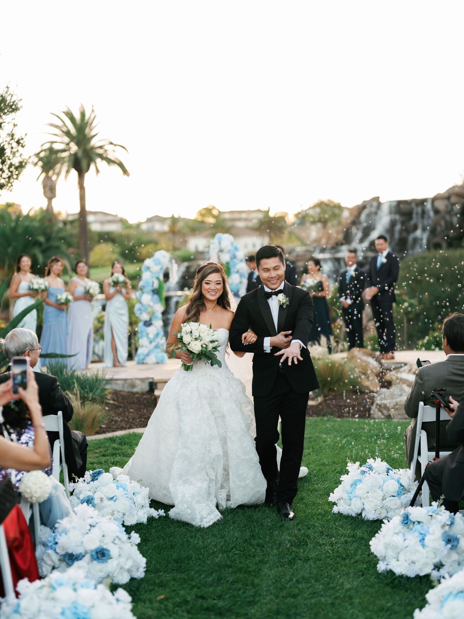 Newlyweds walk down aisle decorated with blue and white flowers as guests celebrate.