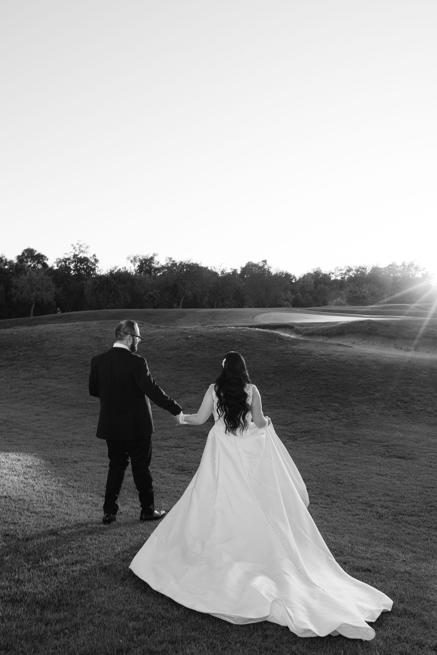 A bride and groom walk hand in hand across a sunlit golf course as her white wedding dress train flows behind them.