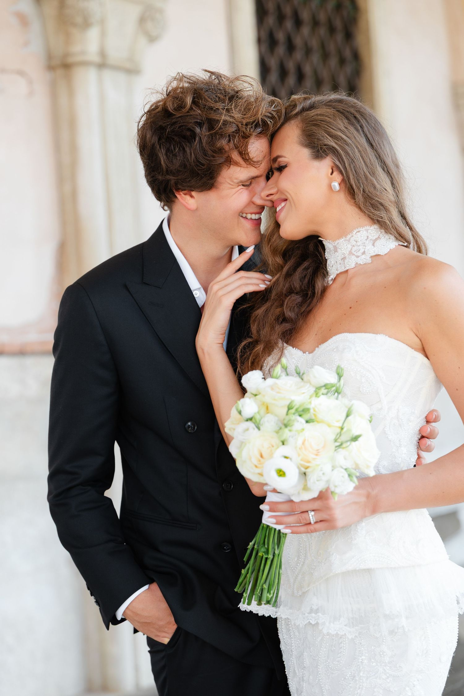 A bride and groom share a romantic moment, smiling forehead-to-forehead, as she holds a white floral bouquet.