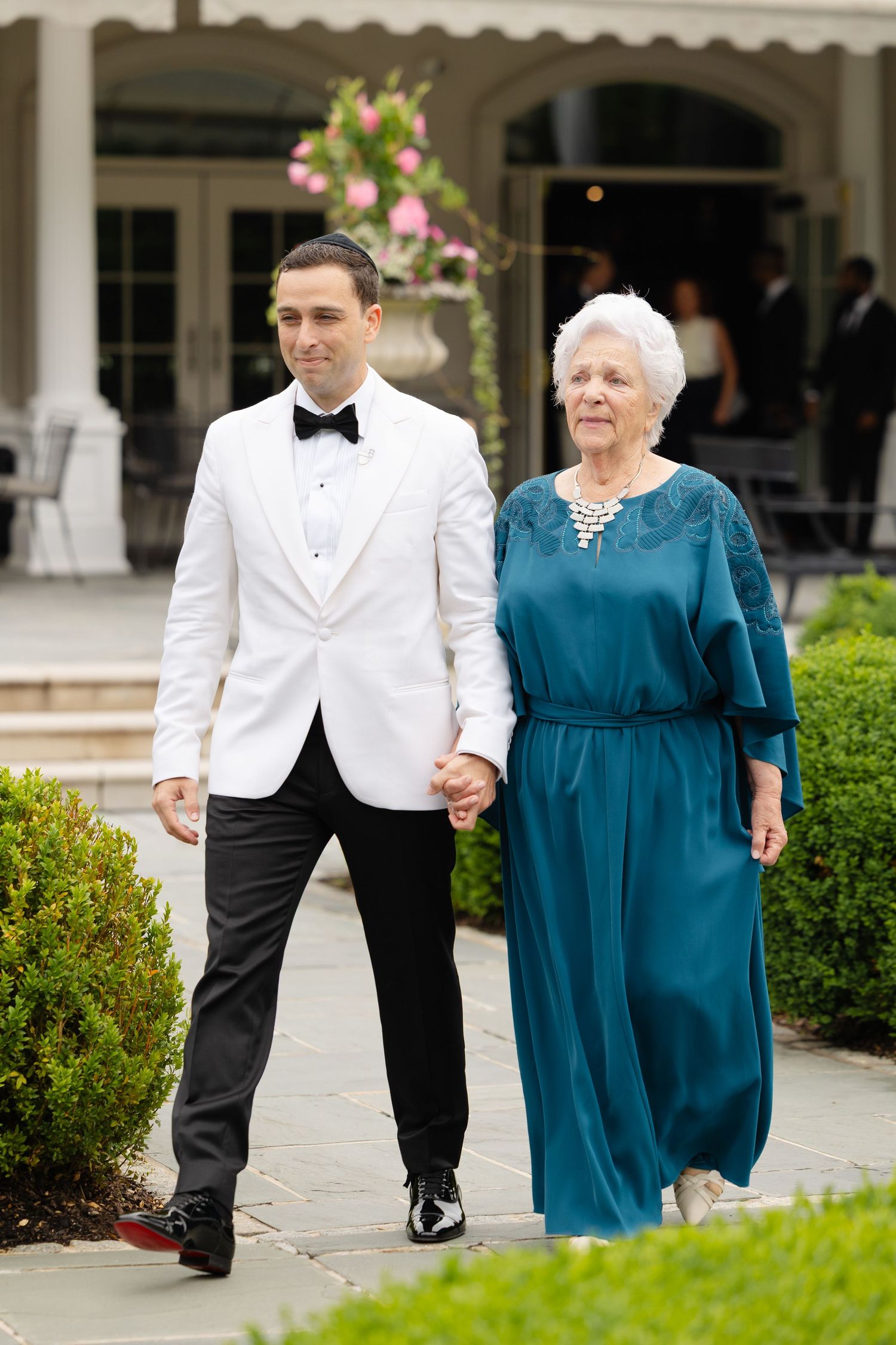 A groom in a white tuxedo jacket escorts an elderly woman in a teal dress along a garden path at a wedding venue.