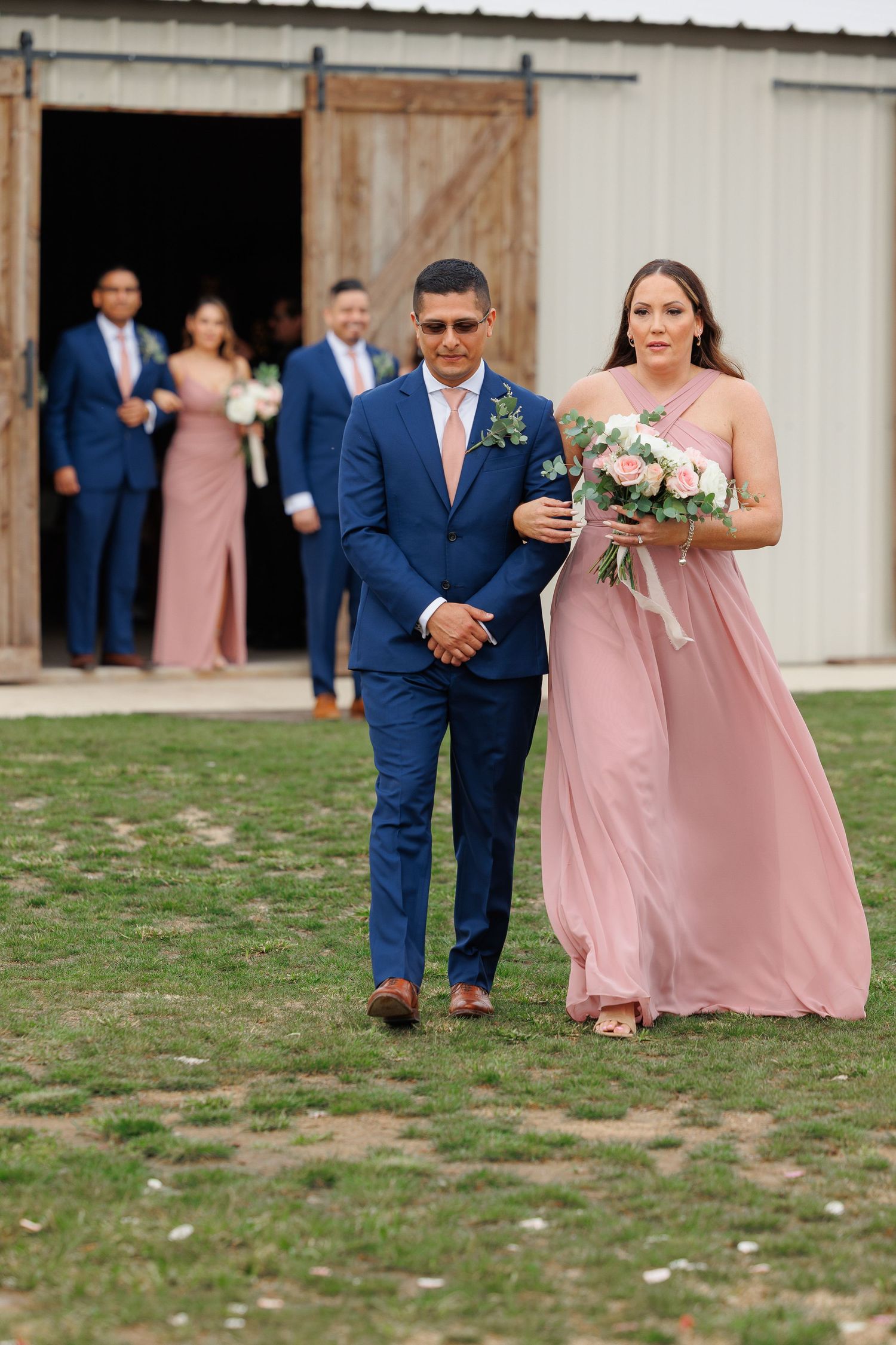 Wedding party members in navy suits and blush pink dresses walk across a grassy field outside a rustic barn venue.