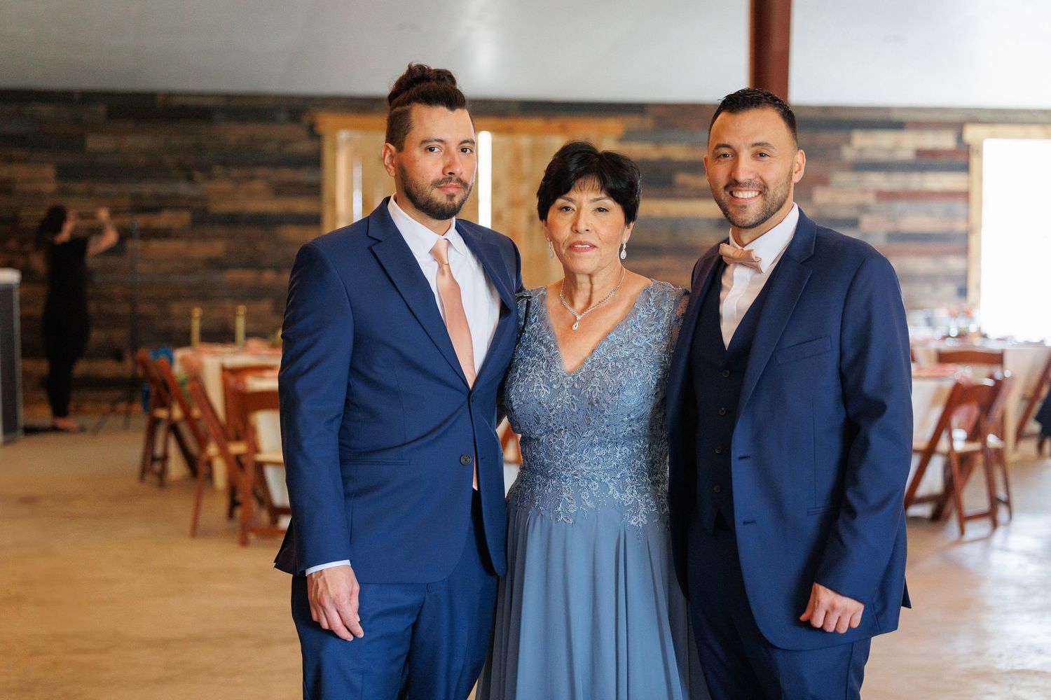 Three people in formal attire pose together at an event venue with a stone wall and wooden chairs in the background.