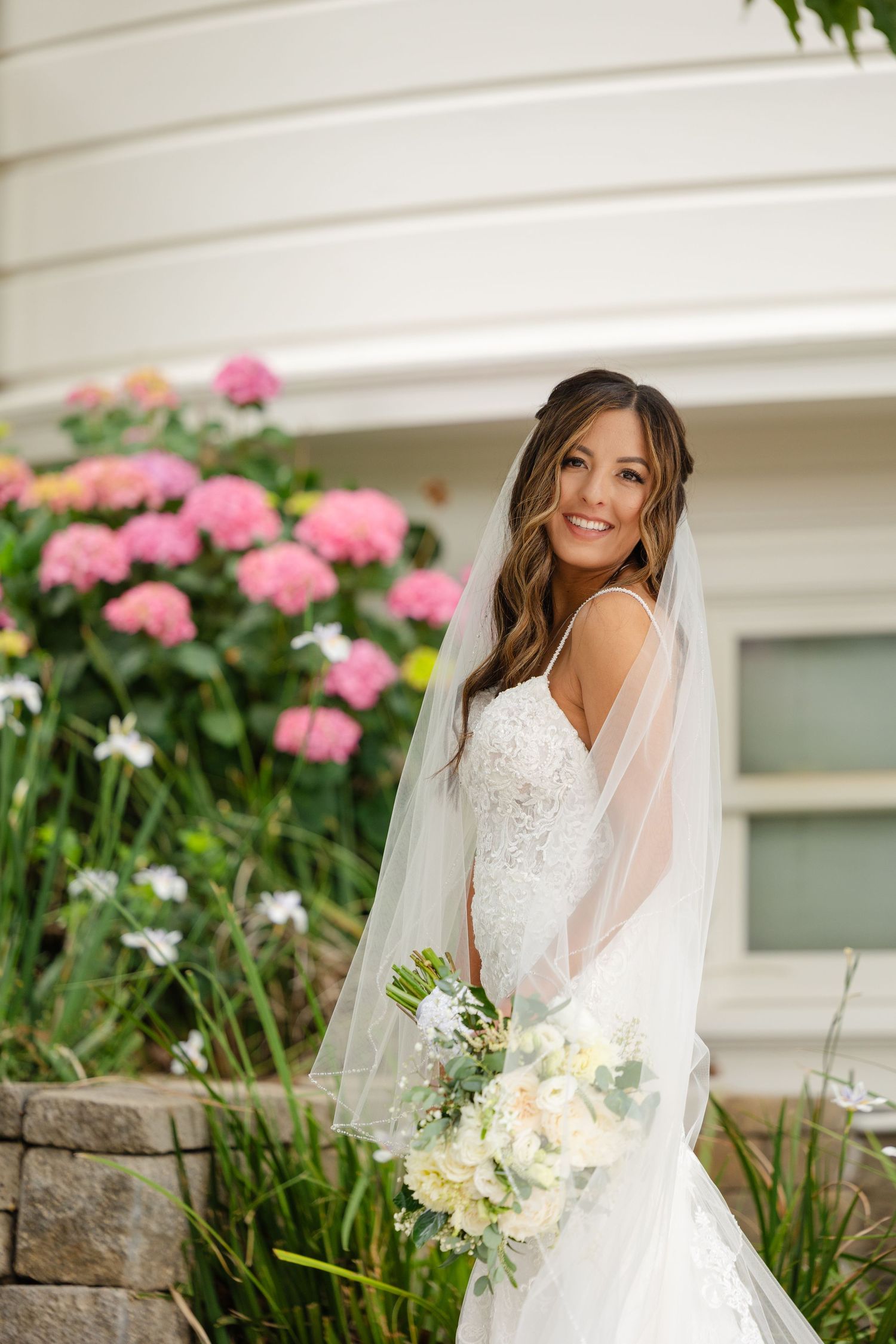 A bride in a white lace wedding dress and veil poses in front of pink flowers and a white wall.