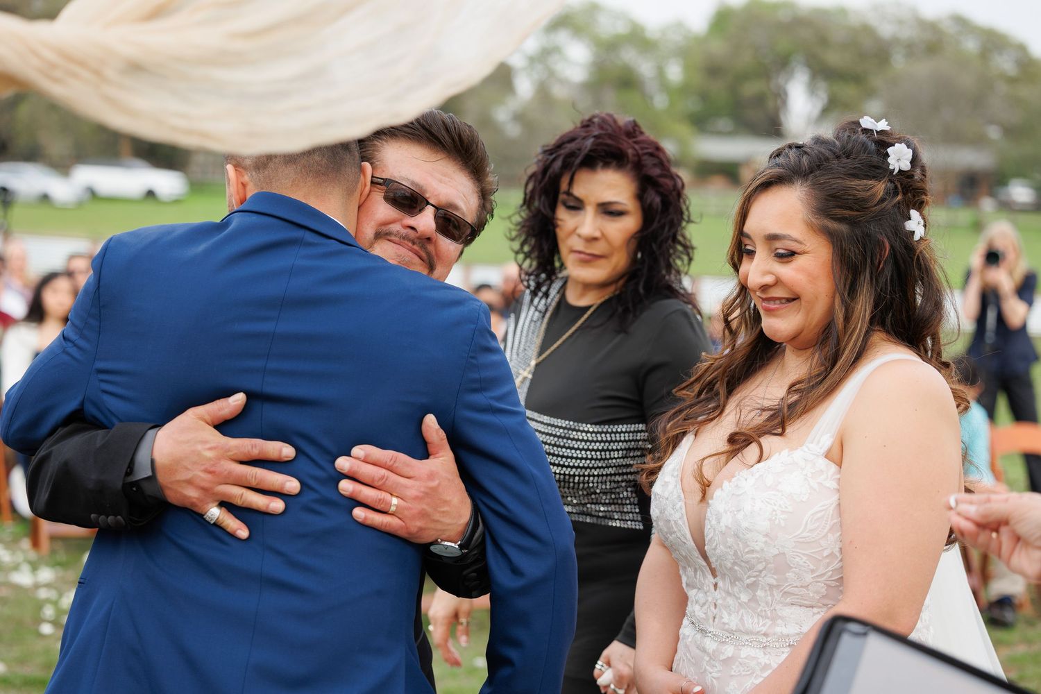 A group of people share an emotional moment at an outdoor wedding celebration on a grassy field.