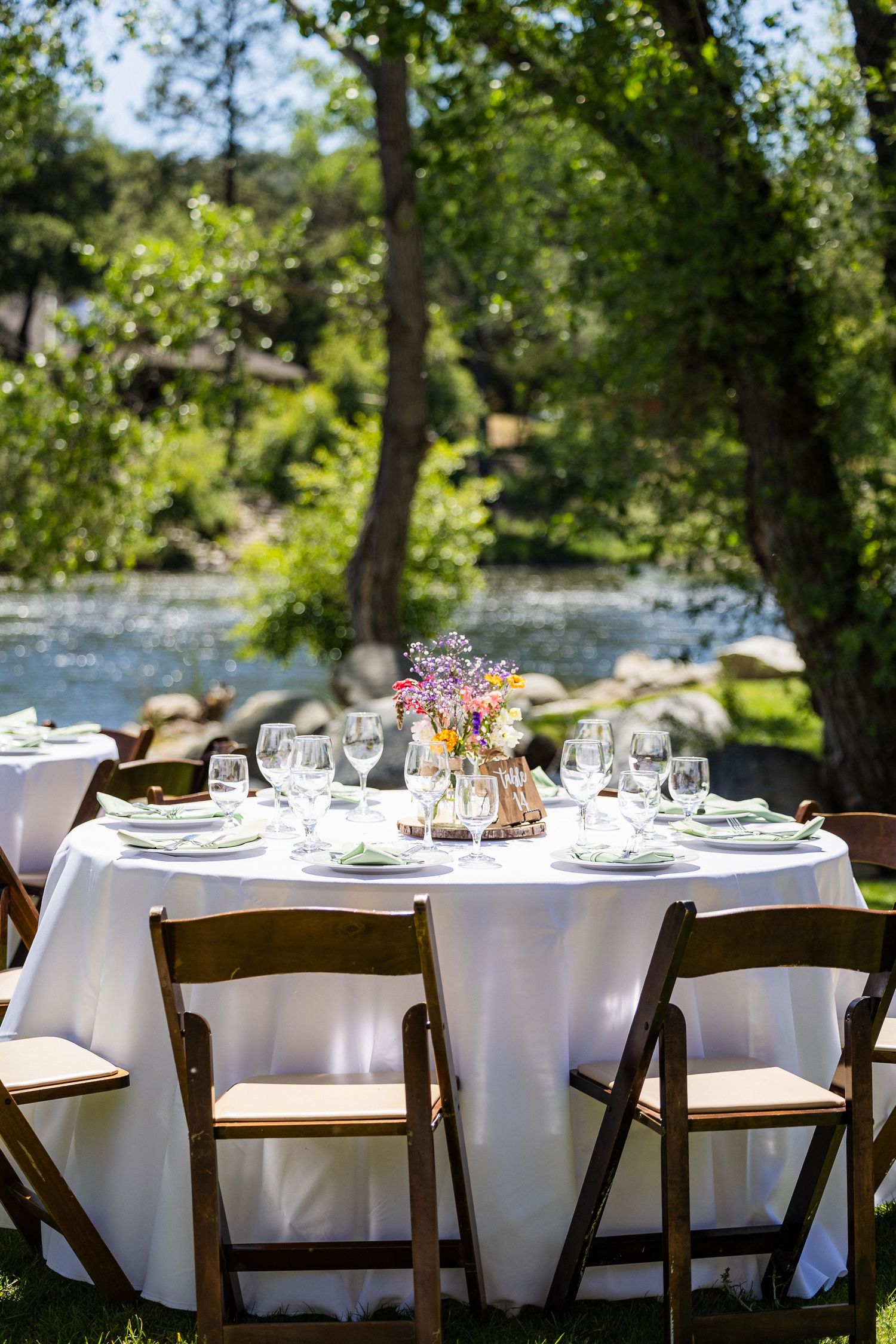 Elegant outdoor dining tables with white tablecloths and place settings arranged under shady trees for a garden event.