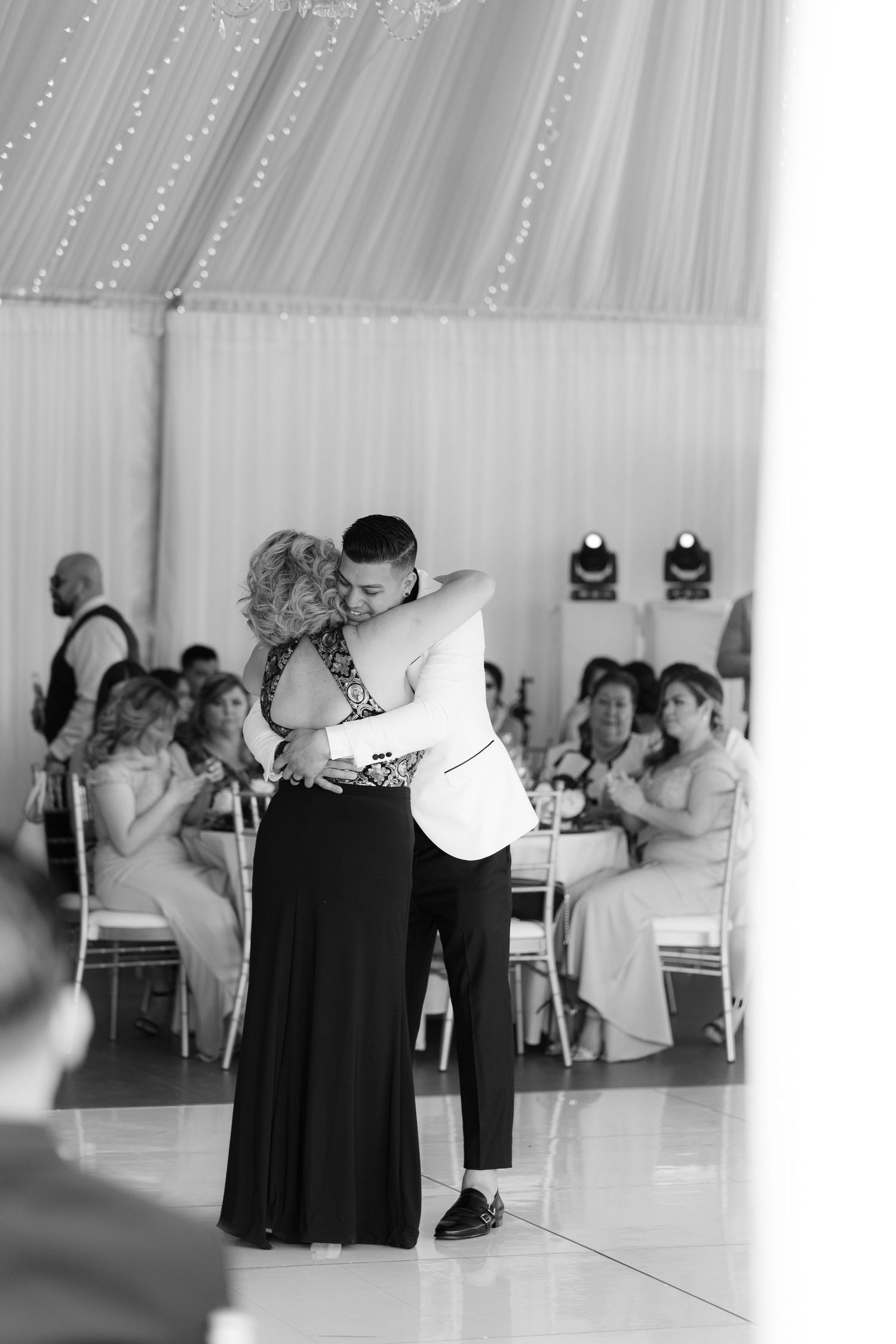 Dancers in formal black and white attire perform together on a dance floor with mirrors and spectators in the background.