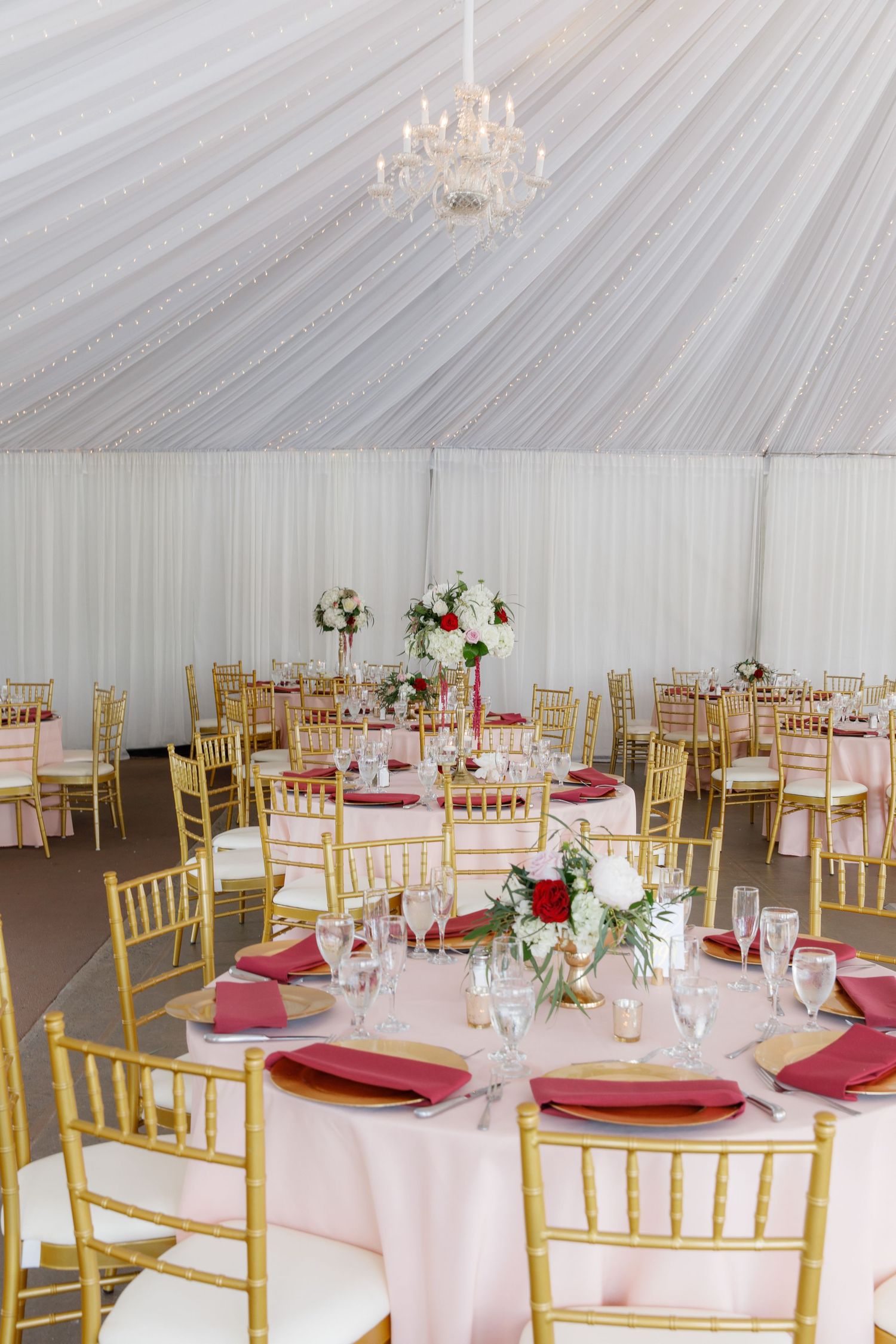 Elegant wedding reception tent with pink and gold decor featuring round tables, chiavari chairs and crystal chandelier.