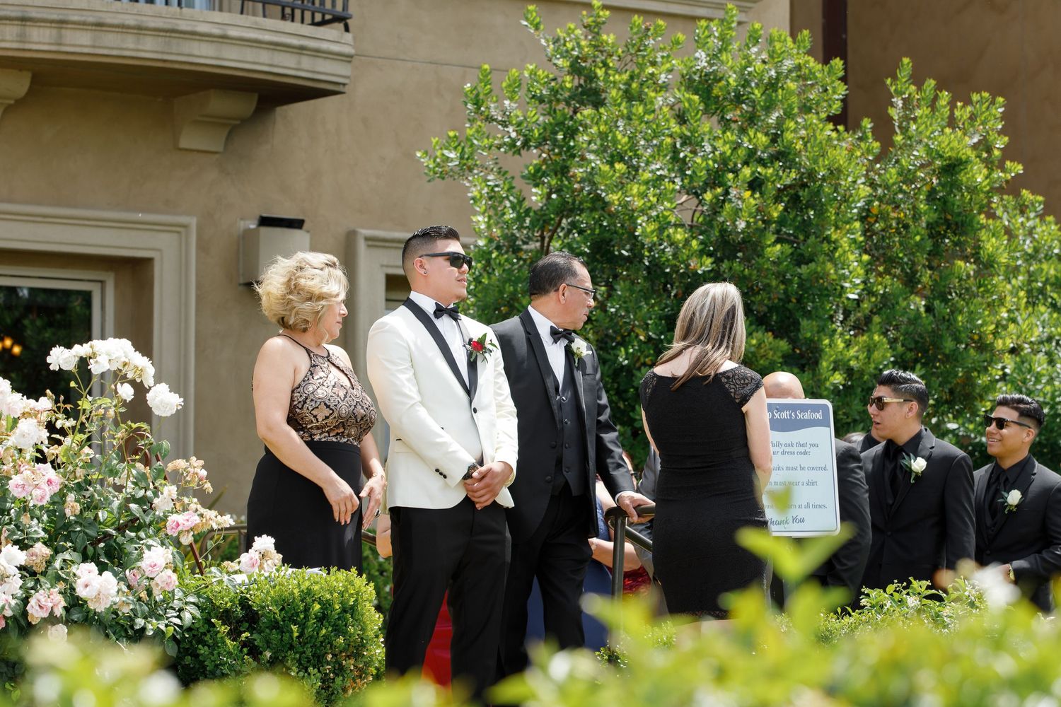 A group of formally dressed people gather outside near flowering bushes and greenery at an elegant event.