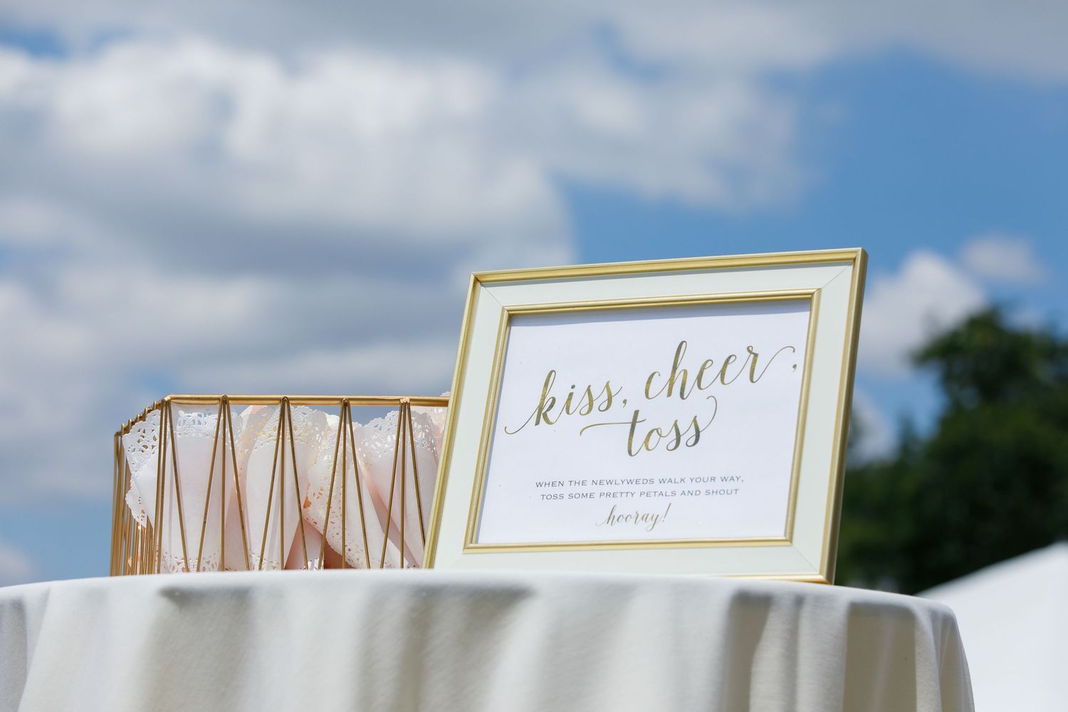 A framed 'kiss cheer less' sign sits beside a rose gold geometric vase on a white tablecloth outdoors.