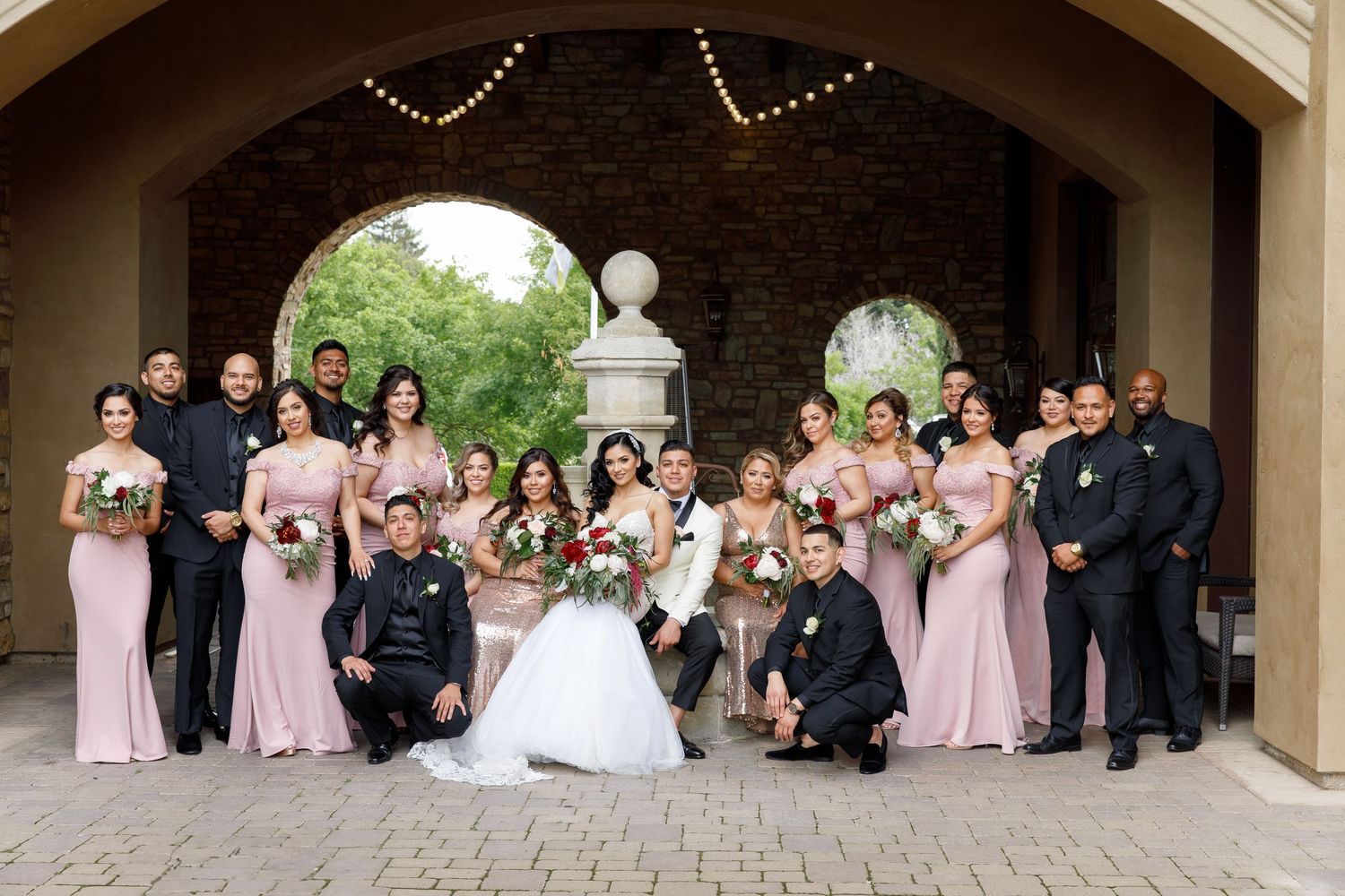 Wedding party in formal attire poses together under covered outdoor walkway with stone columns.