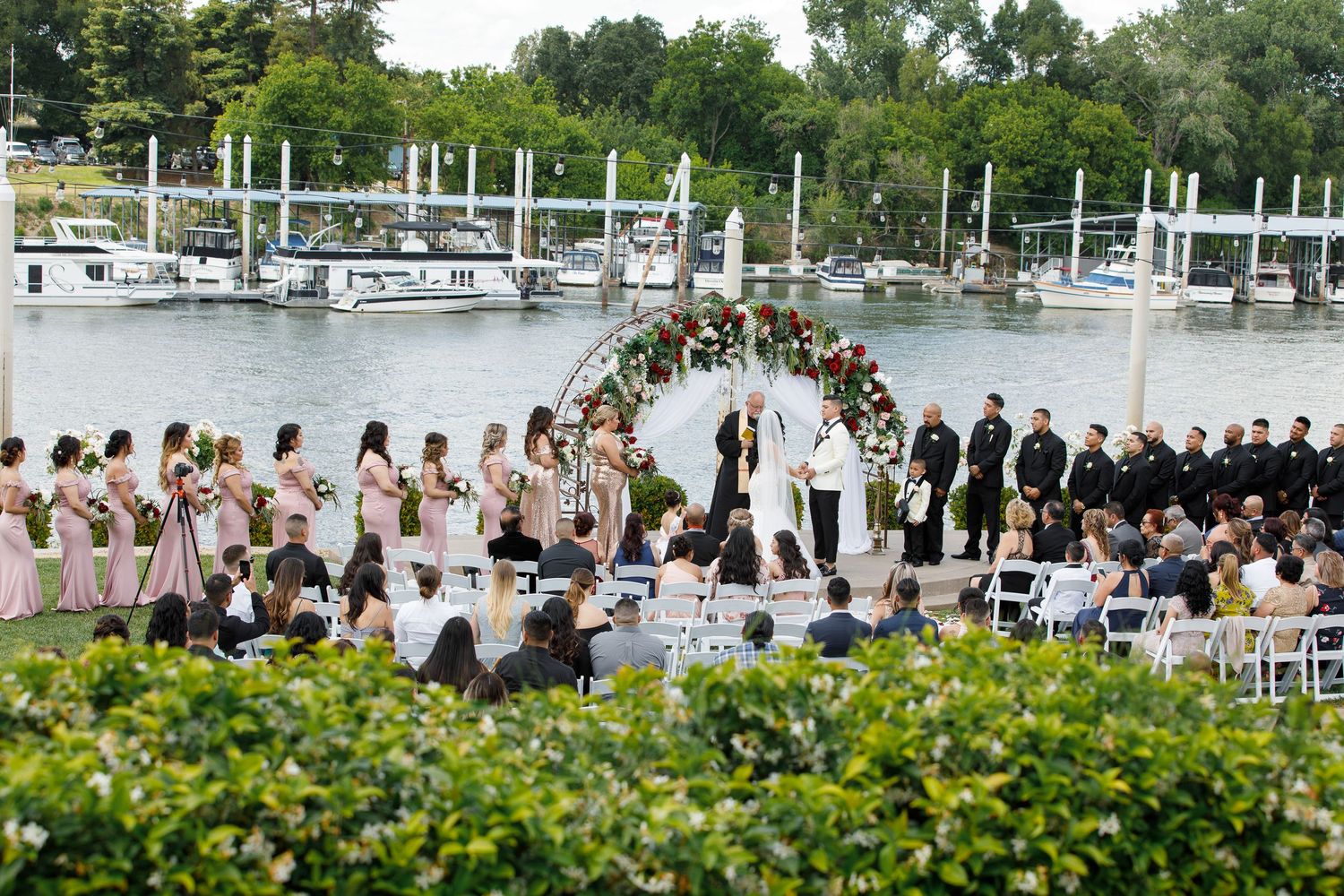 Wedding guests gather outdoors near a marina with boats and greenery for a waterfront ceremony.
