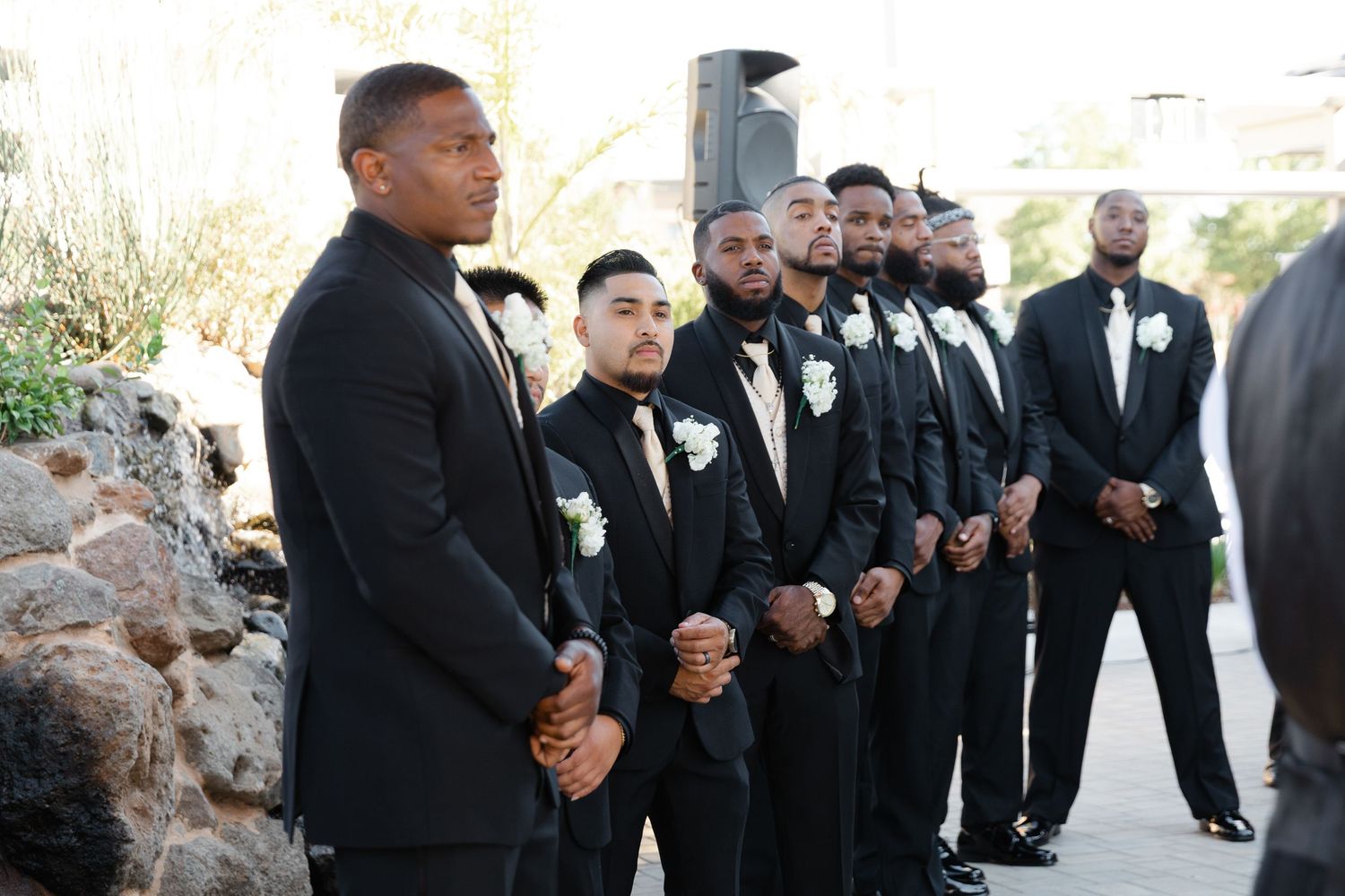 A group of groomsmen in black tuxedos stand together outdoors during a wedding ceremony.