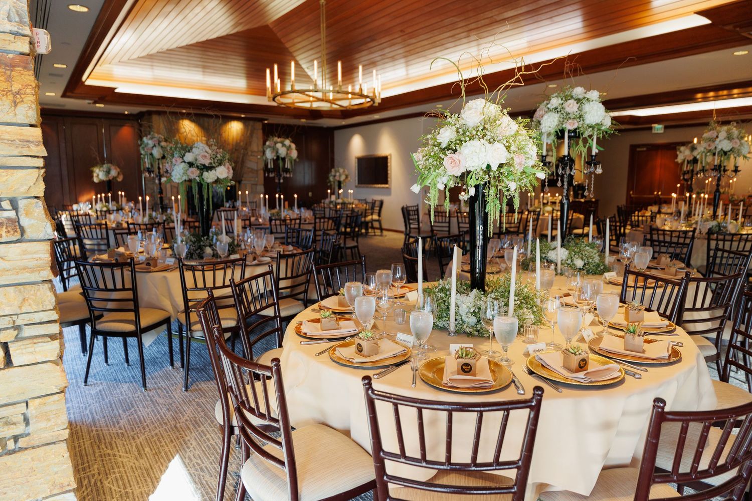 Elegant rustic wedding reception venue with dark wood tables, chiavari chairs, and floral centerpieces under warm lighting.