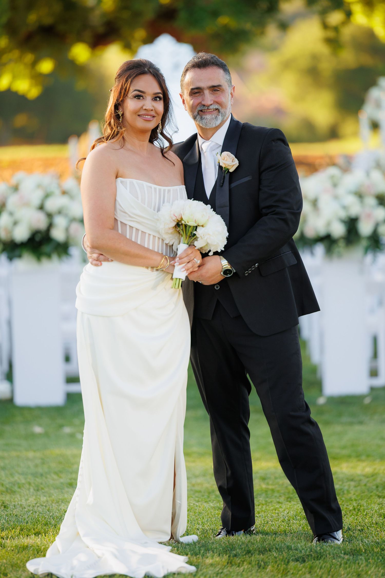 Newlywed couple poses together on a sunny lawn with white chairs and floral decorations in the background.