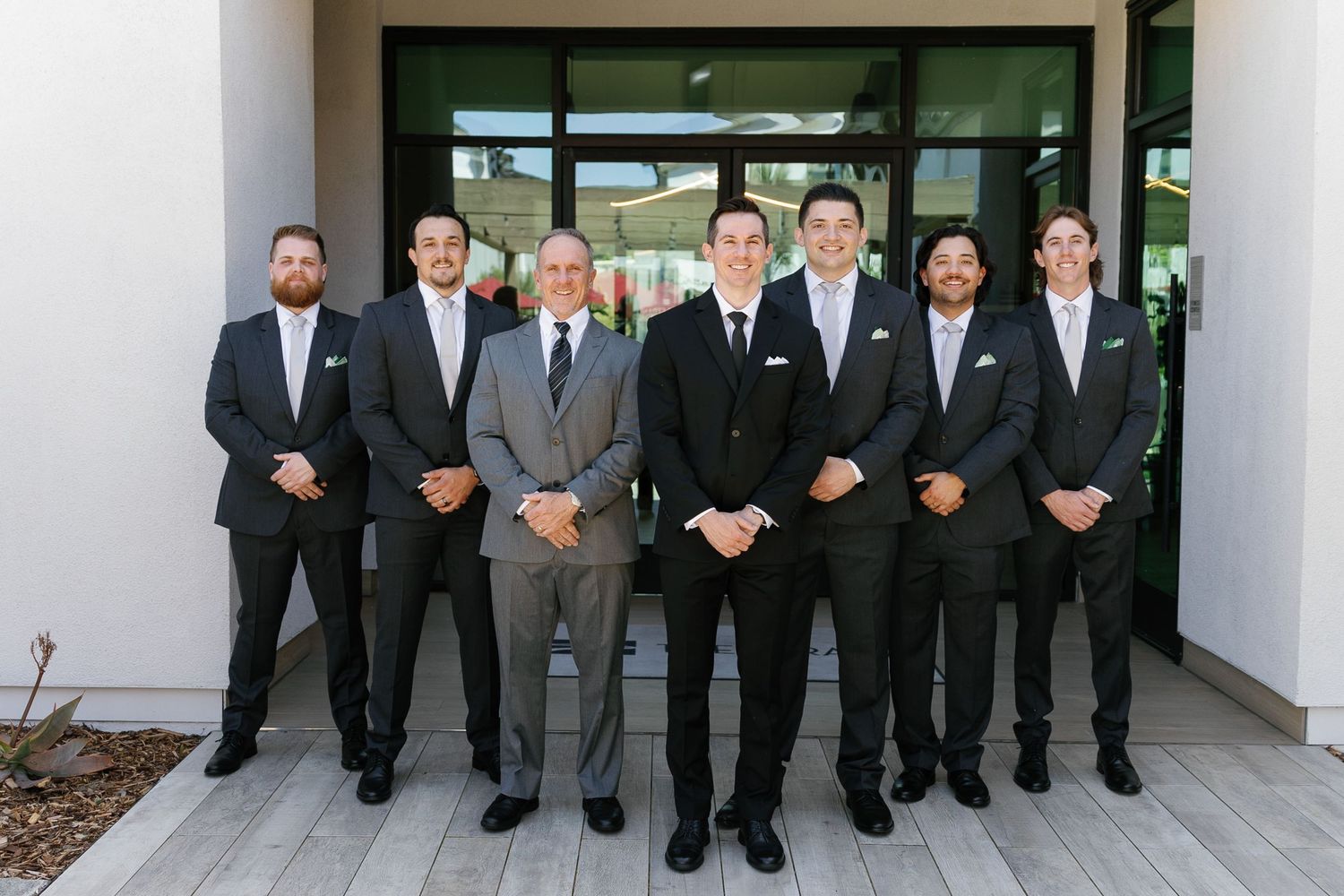 A group of men in formal black suits and ties stand together in a row outside a building entrance.