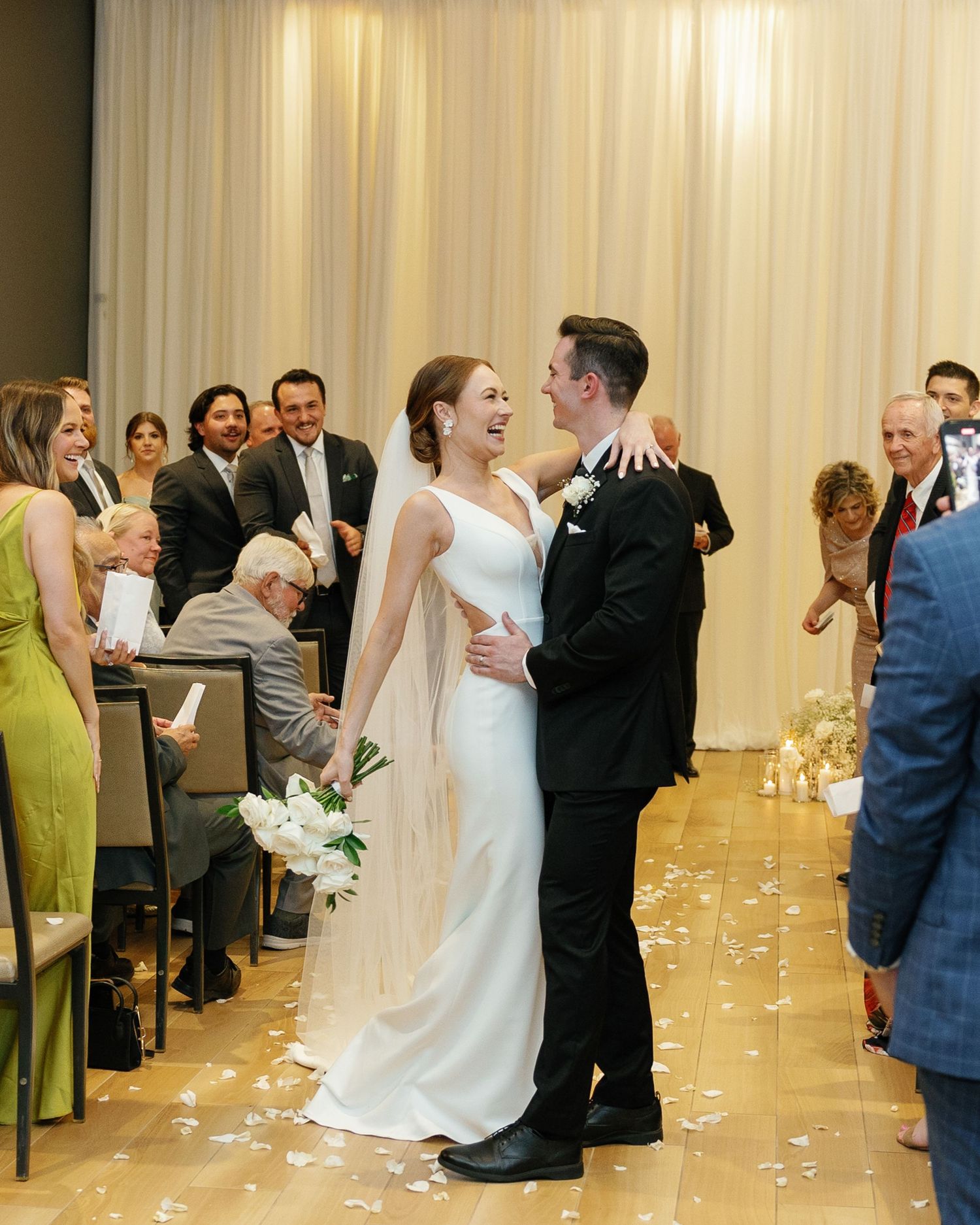 A couple shares their first dance at a wedding reception with guests looking on and flower petals scattered on the floor.