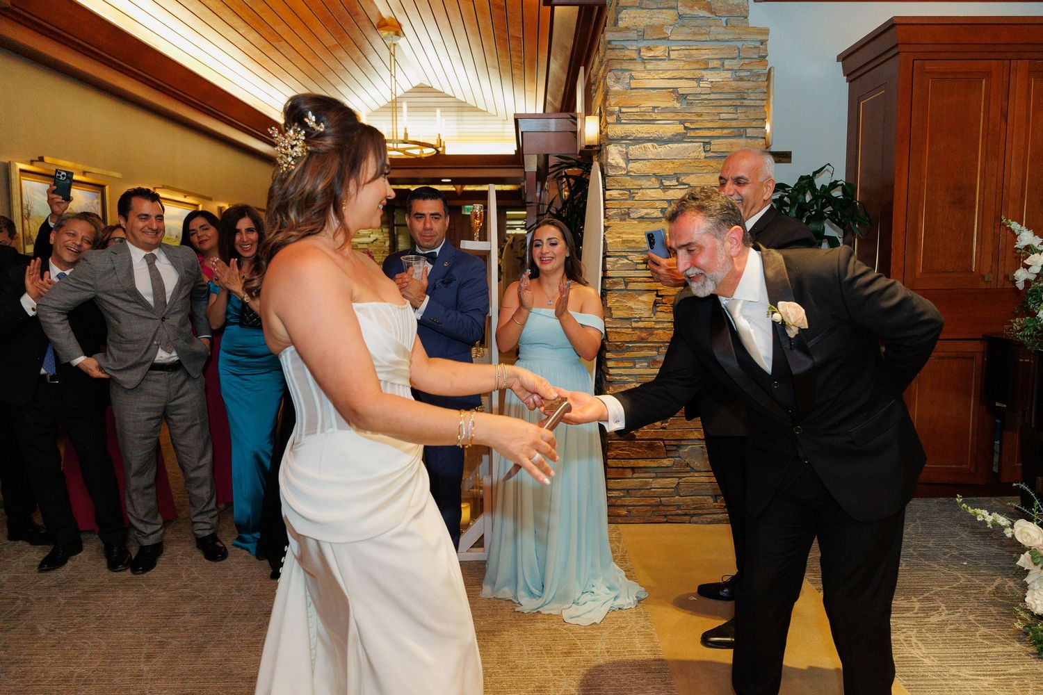 A wedding celebration shows a dance moment at a reception venue with a stone wall and wooden ceiling.