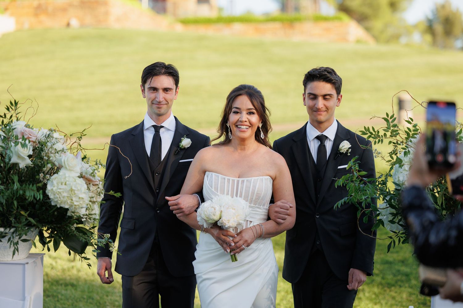 Wedding party walks together through a sunny garden setting during an outdoor celebration.