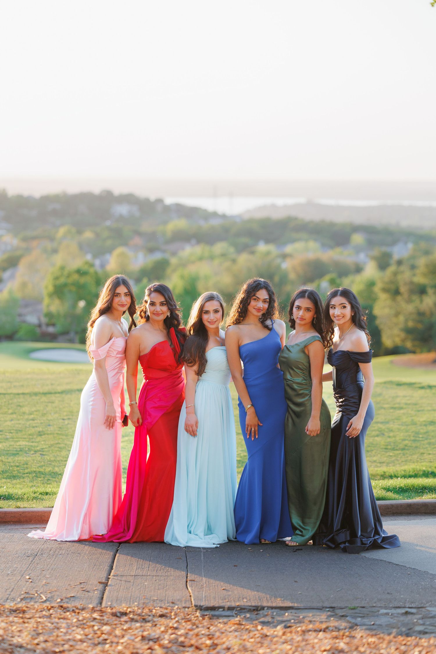A group of friends in formal dresses pose together against a scenic outdoor backdrop at sunset.