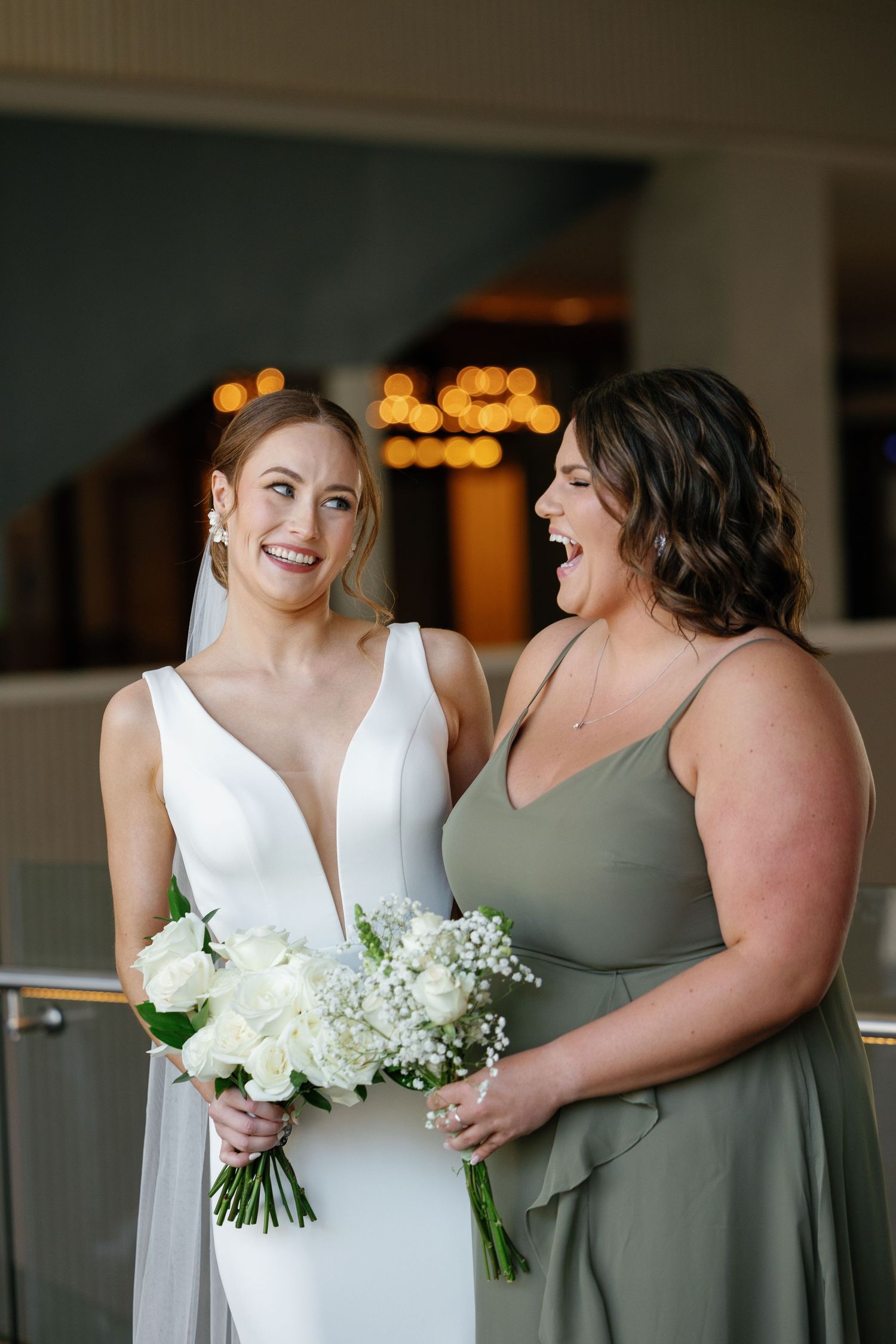 Candid wedding party moment showing bridesmaids in sage green dresses laughing together while holding white bouquets.
