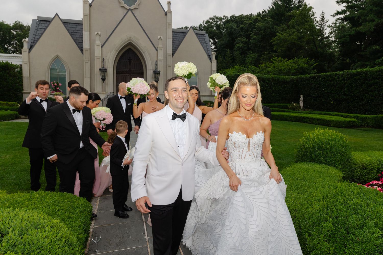 Wedding party walks across a lush green lawn outside a white church steeple on a summer afternoon.