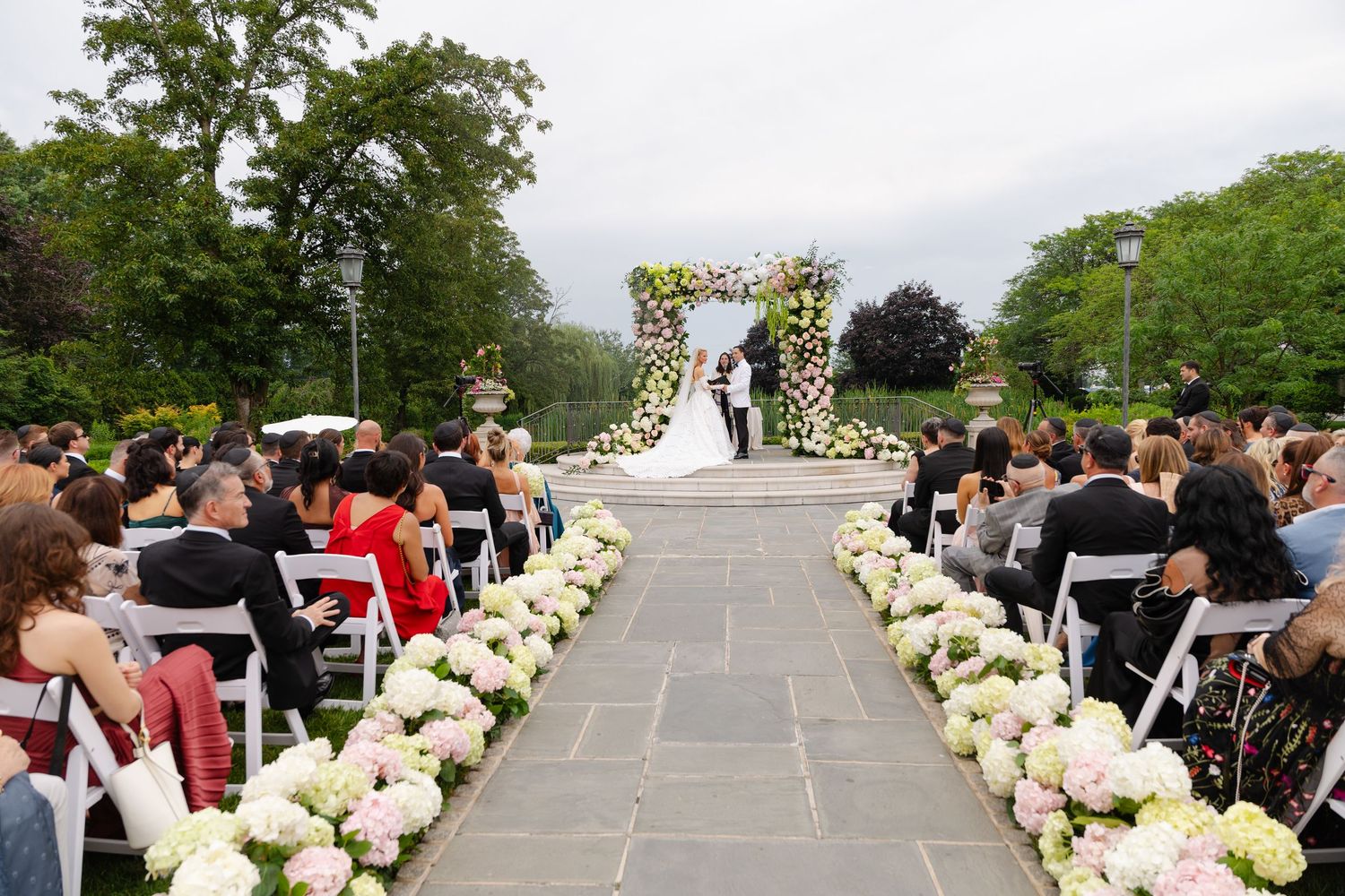 Outdoor wedding ceremony setup with white floral arch and aisle lined with white roses on stone patio surrounded by trees.