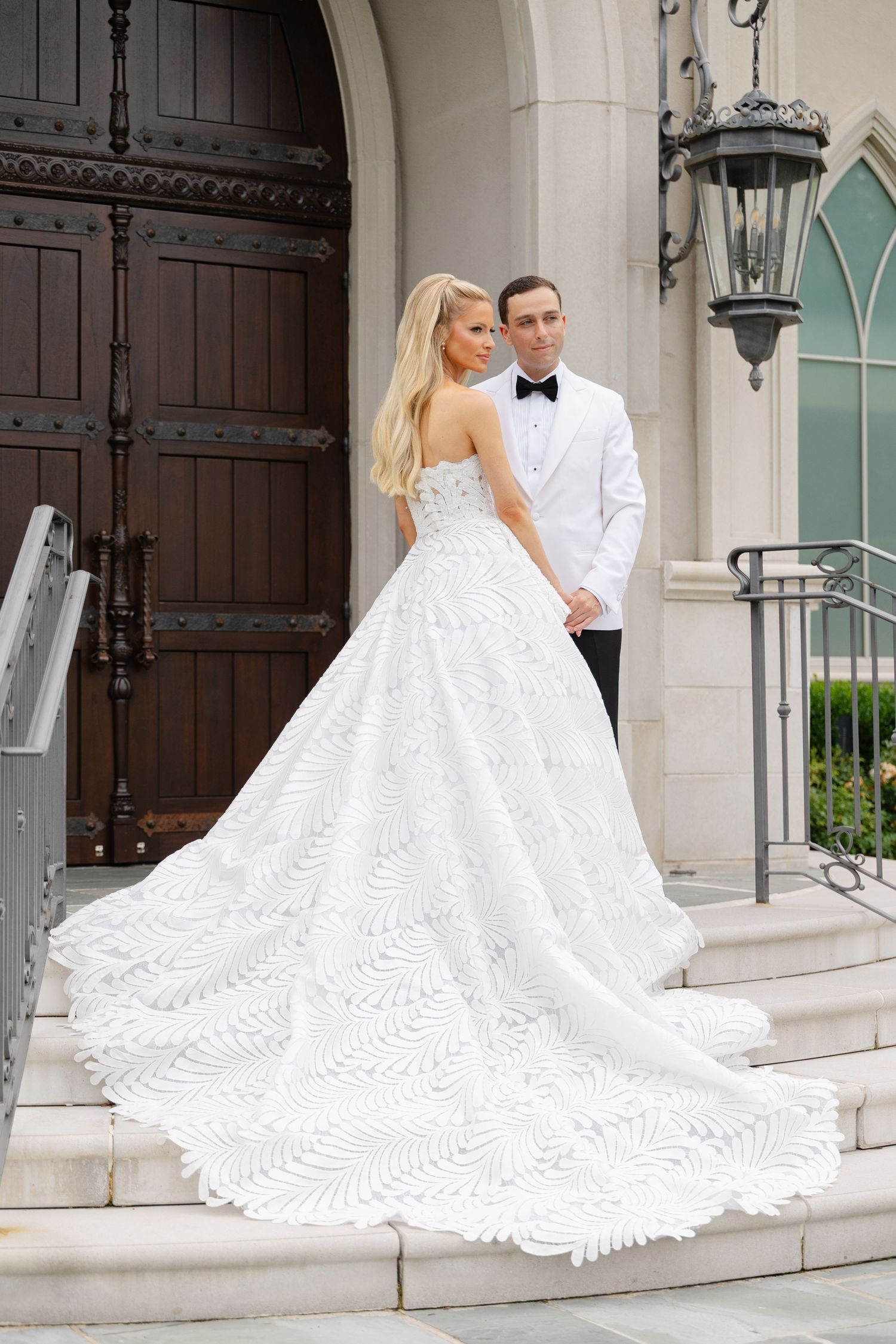 A couple in wedding attire poses for photos on the steps of an elegant stone building with ornate wooden doors.
