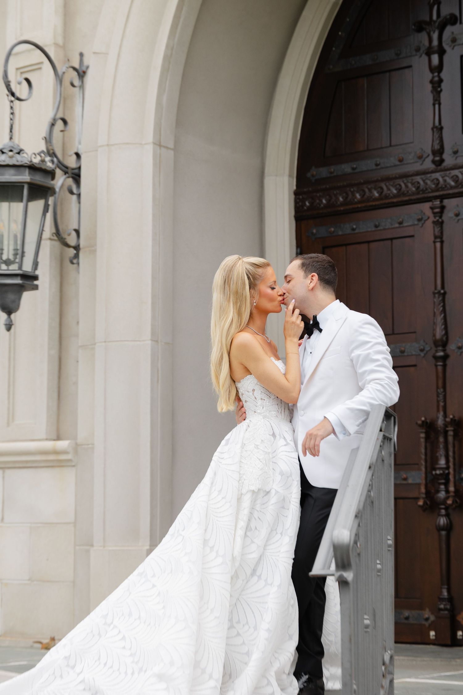A couple in elegant wedding attire embrace near an ornate wooden door with a vintage lantern on a stone archway.