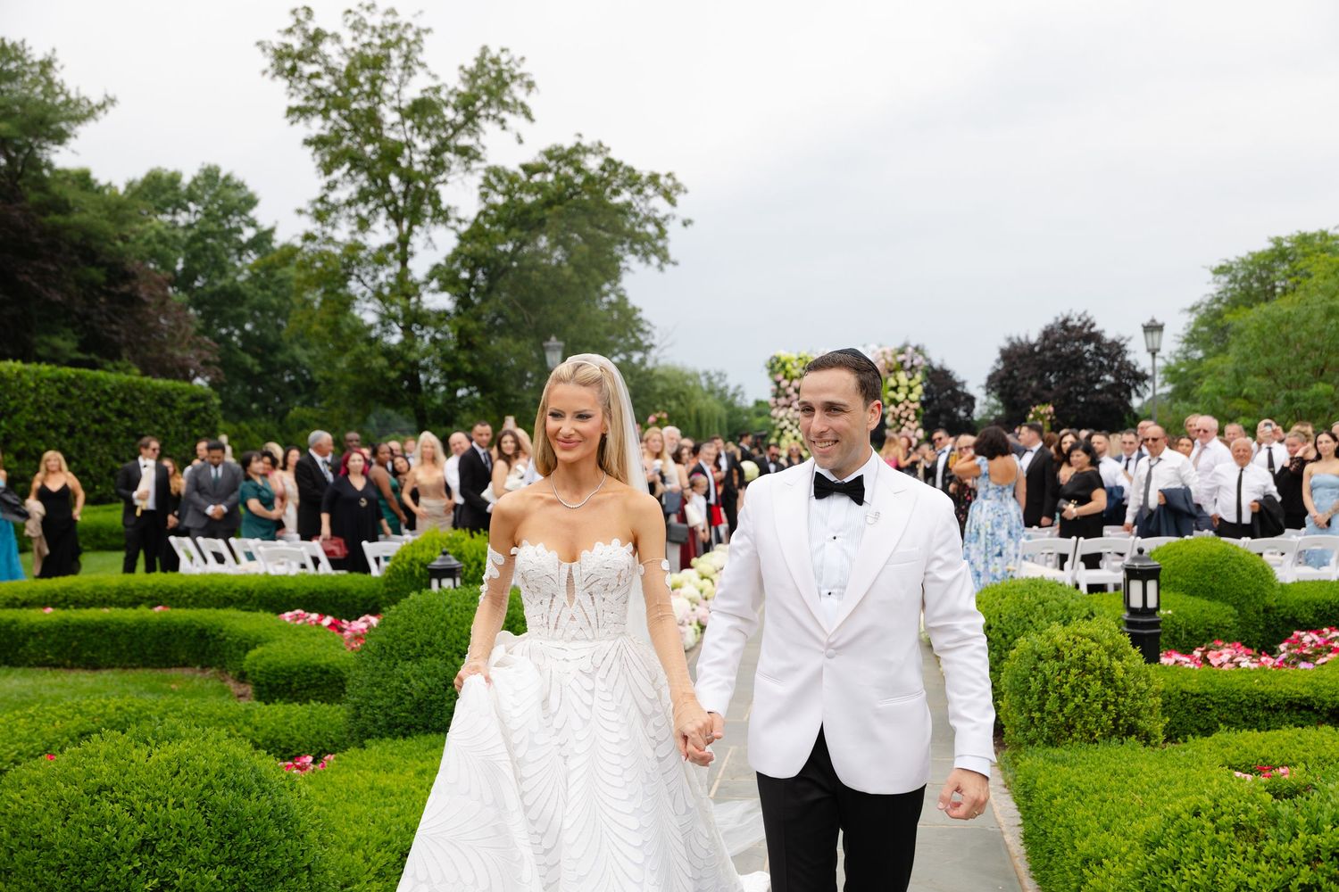 A wedding party celebrates outdoors on a lush green lawn surrounded by trees and guests.