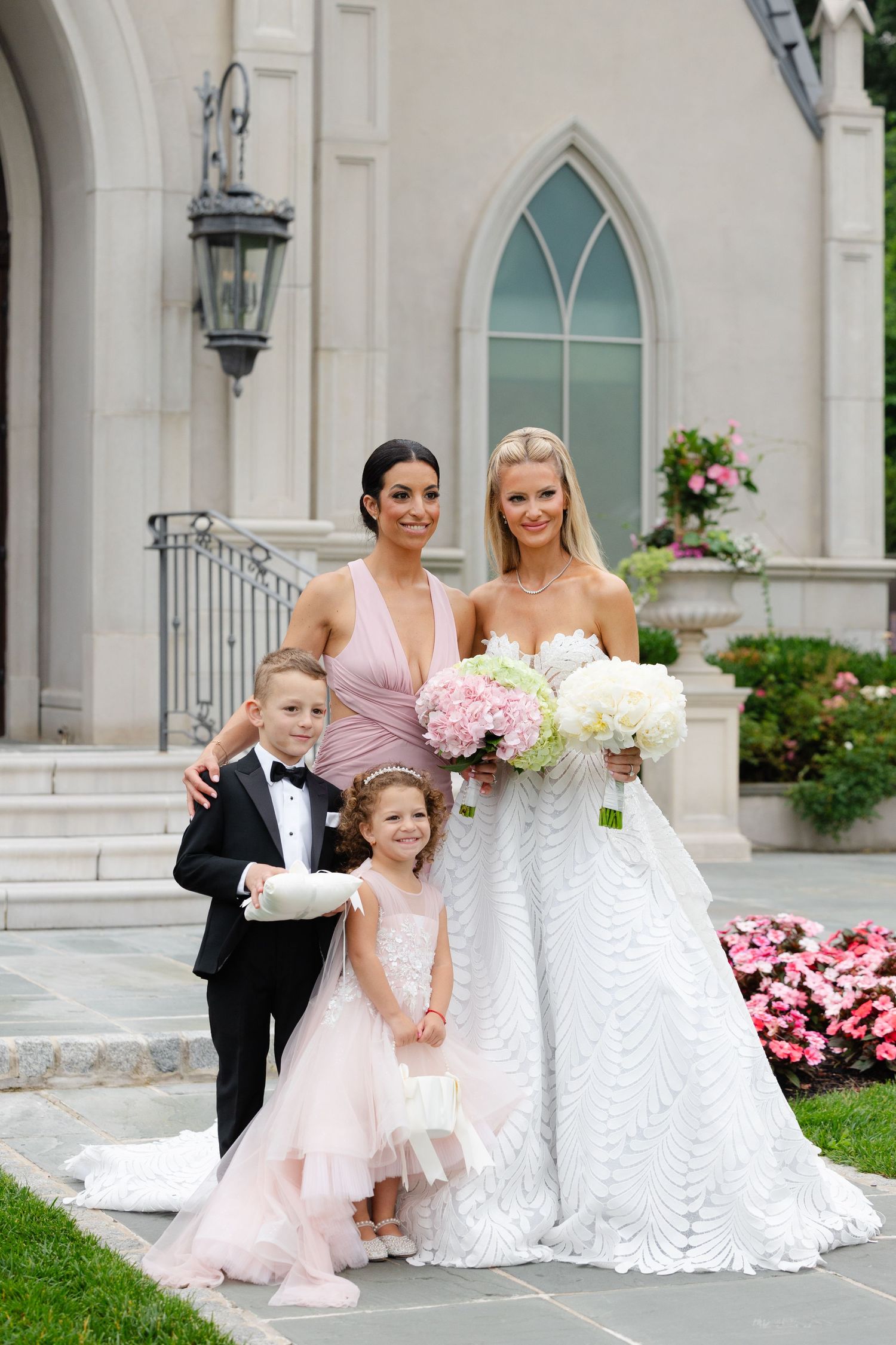Wedding party poses in front of church with white gowns and pink bridesmaid dresses amid blooming flowers.