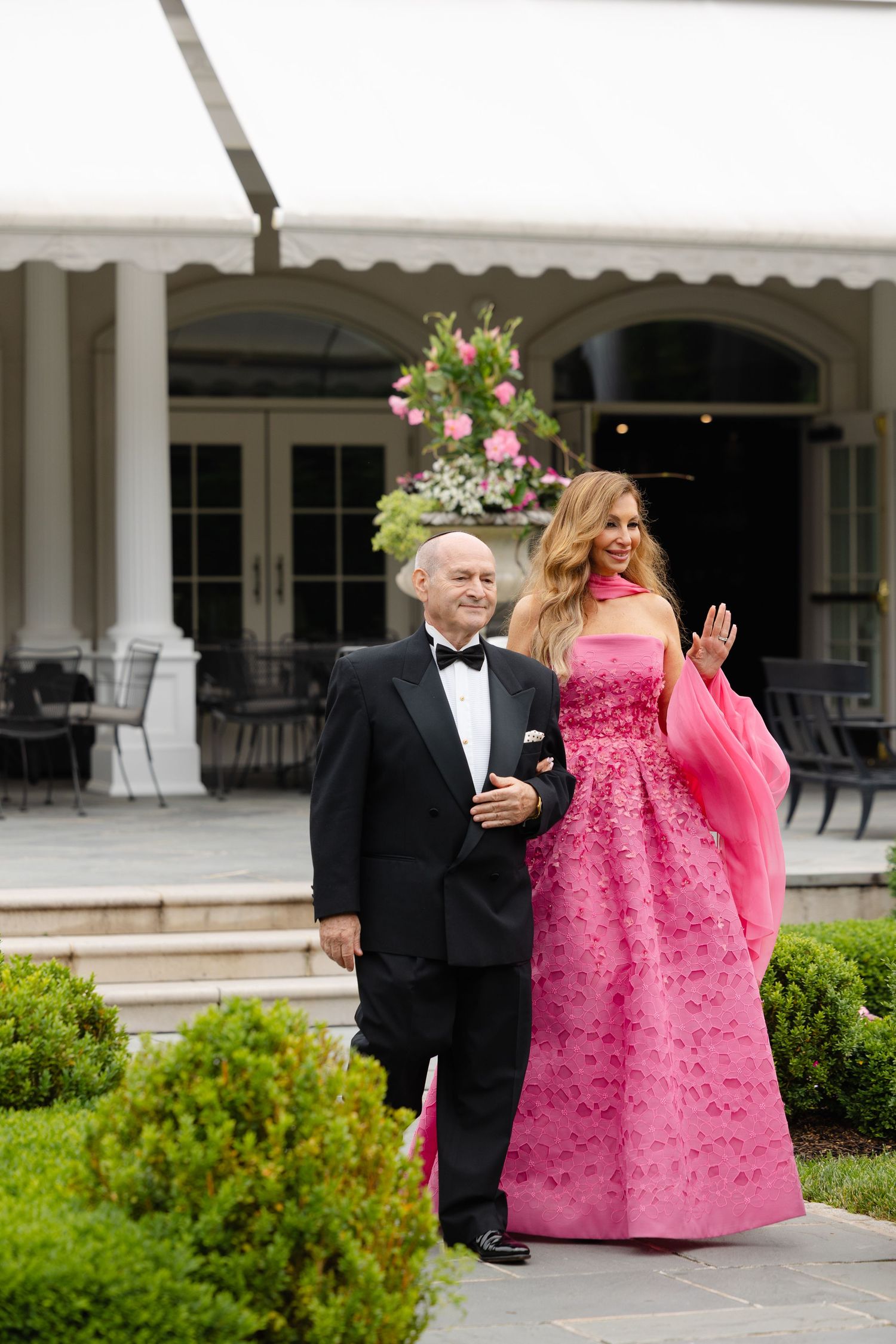 Two people walking outdoors, with one wearing a bright pink voluminous gown and the other in a formal black tuxedo.