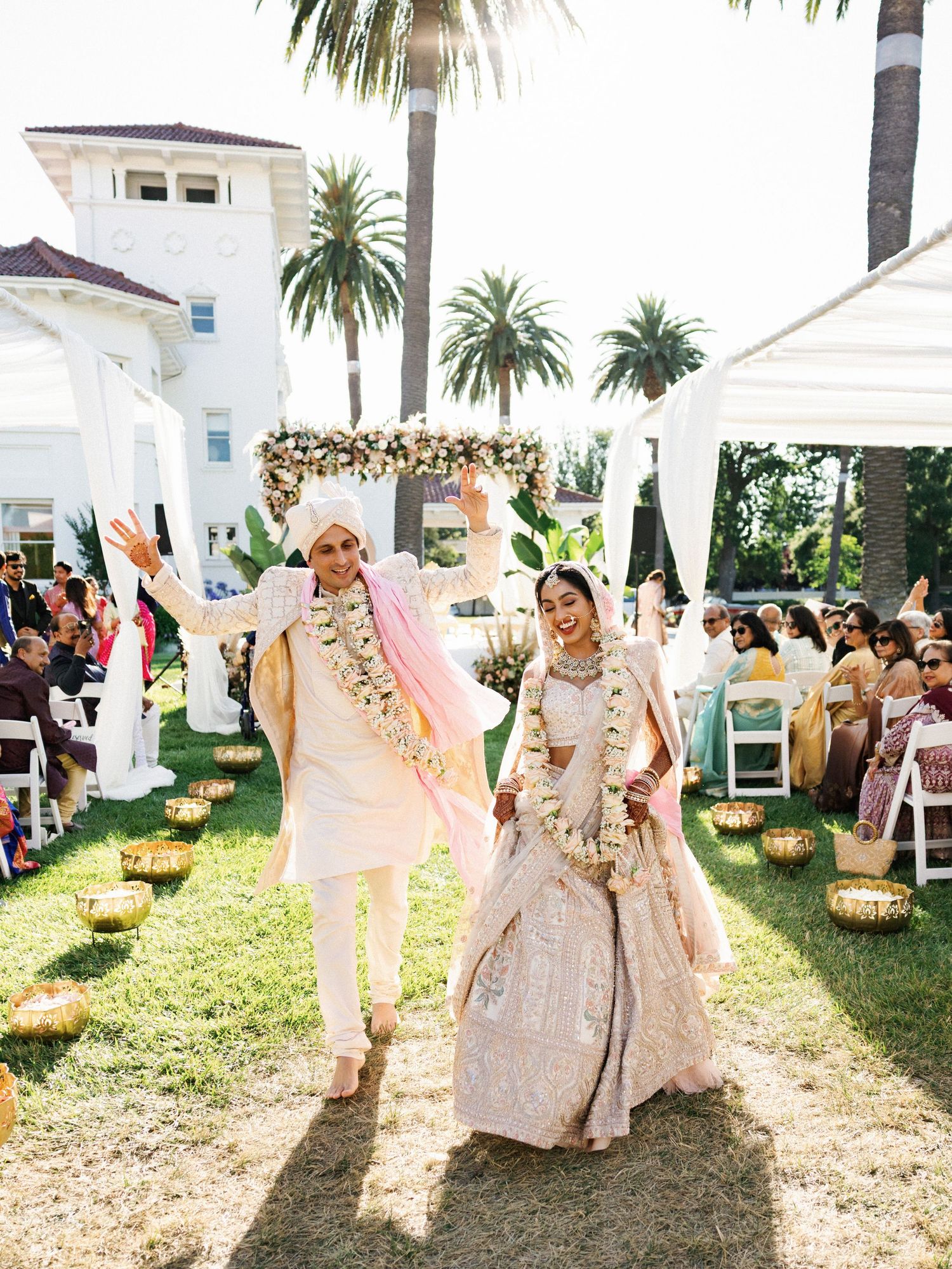 Joyful Indian bride and groom in traditional attire celebrate after outdoor wedding ceremony with palm trees and white venue.