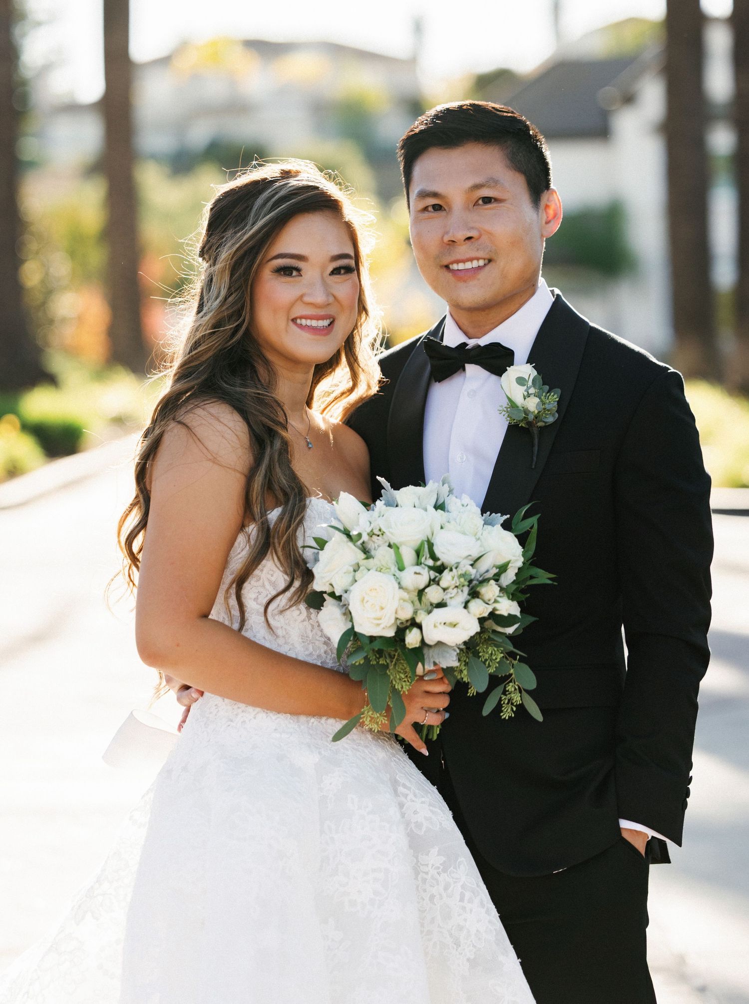 Bride and groom posing outdoors, bride holding white rose bouquet in lace gown, groom in black tuxedo with bow tie.
