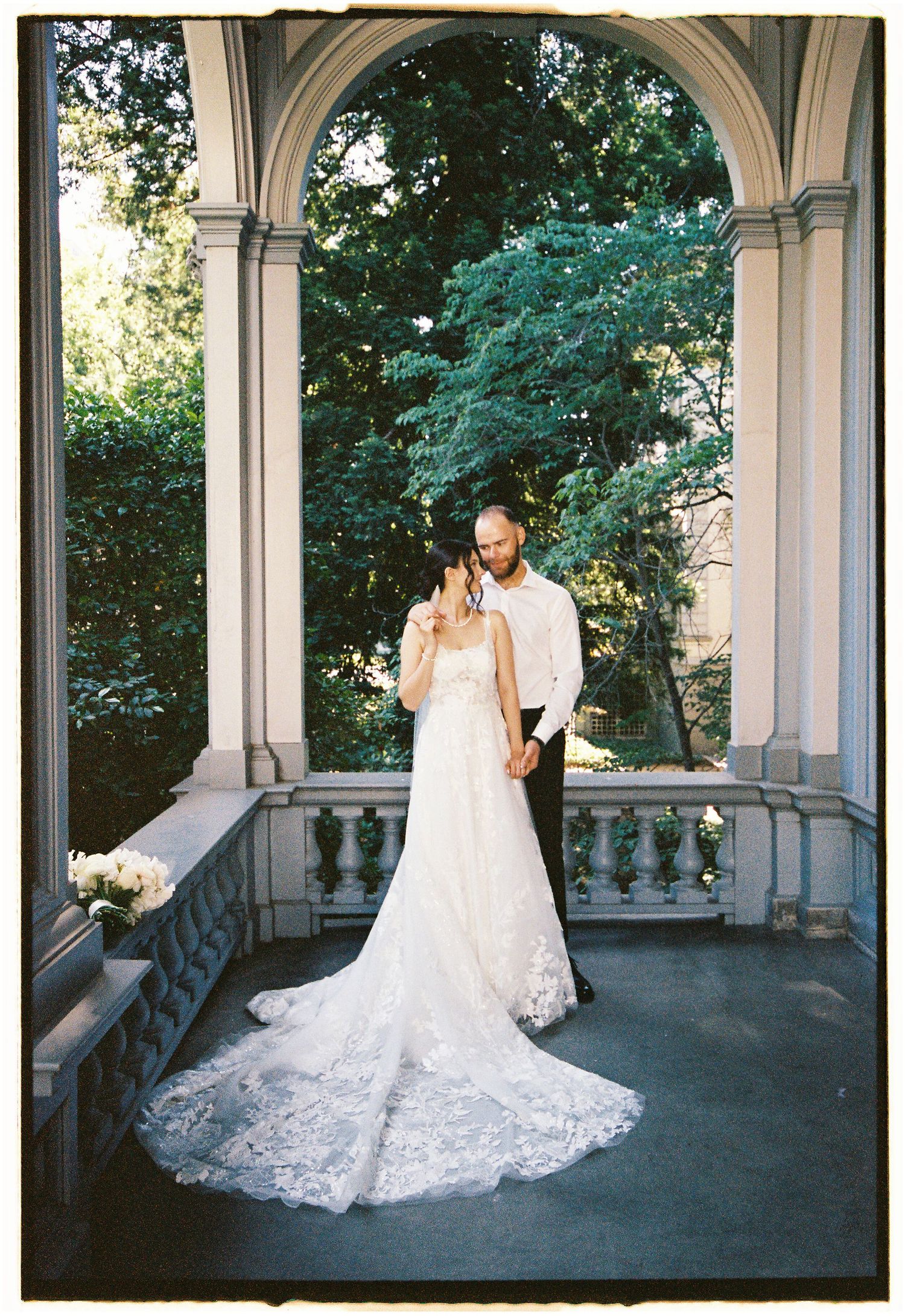 Bride and groom share a romantic moment on an elegant stone balcony with arched columns and lush green trees.