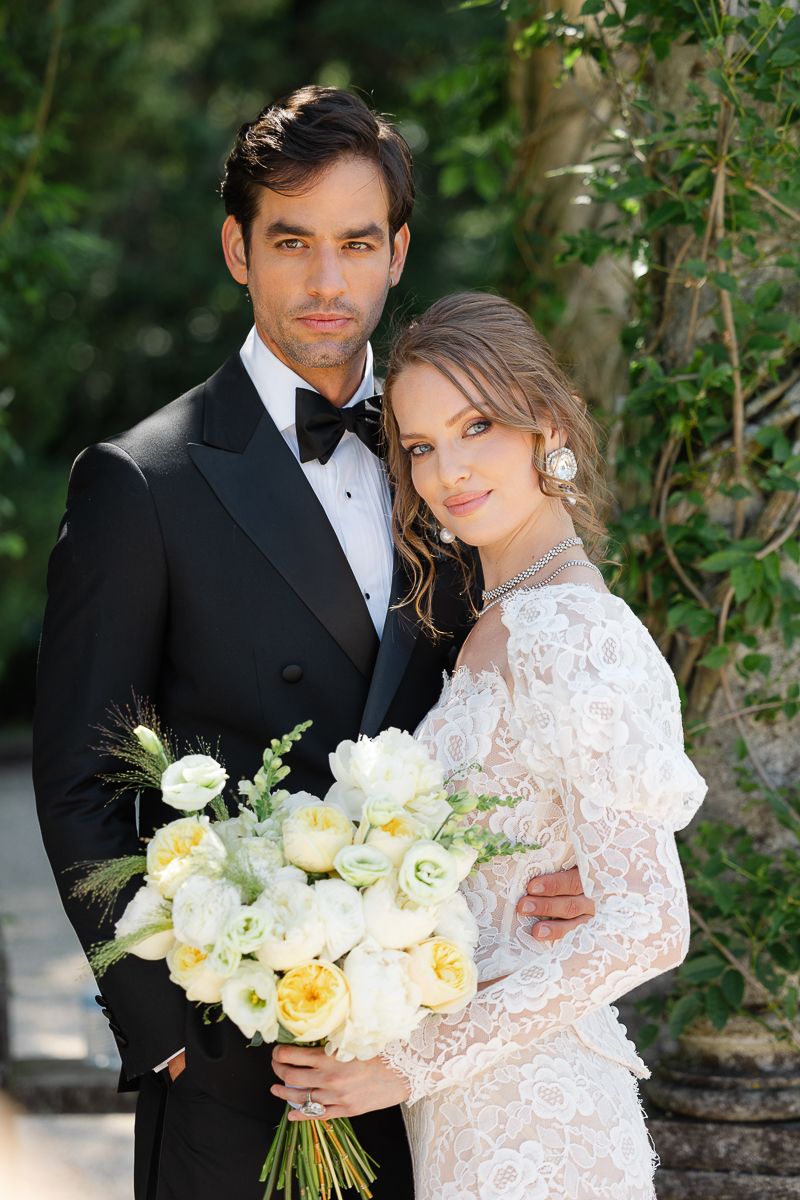 Elegant couple in formal wedding attire pose with yellow and white bouquet against garden backdrop.