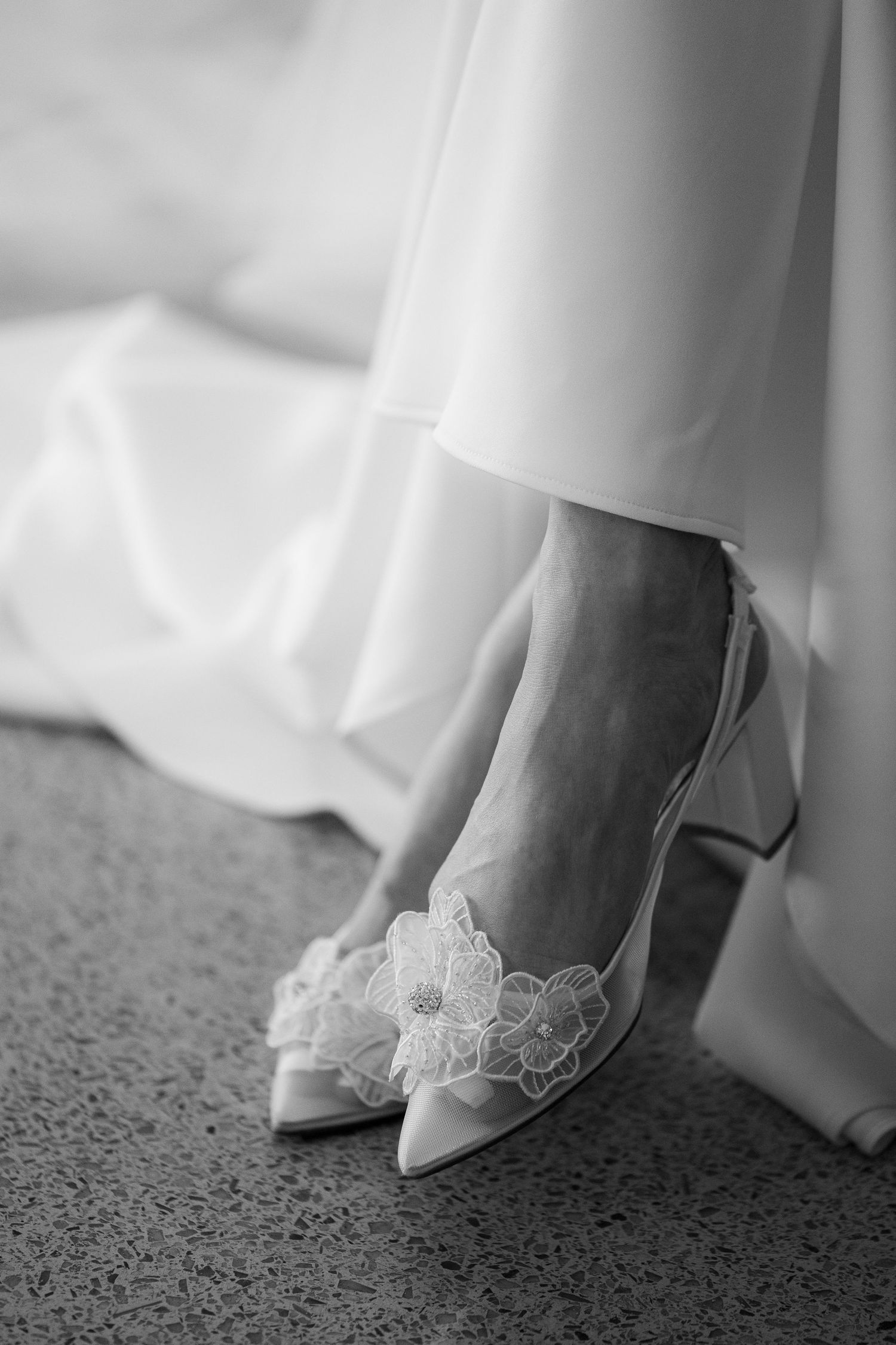A close-up view of elegant white lace bridal shoes on gray carpet, showing delicate floral details.