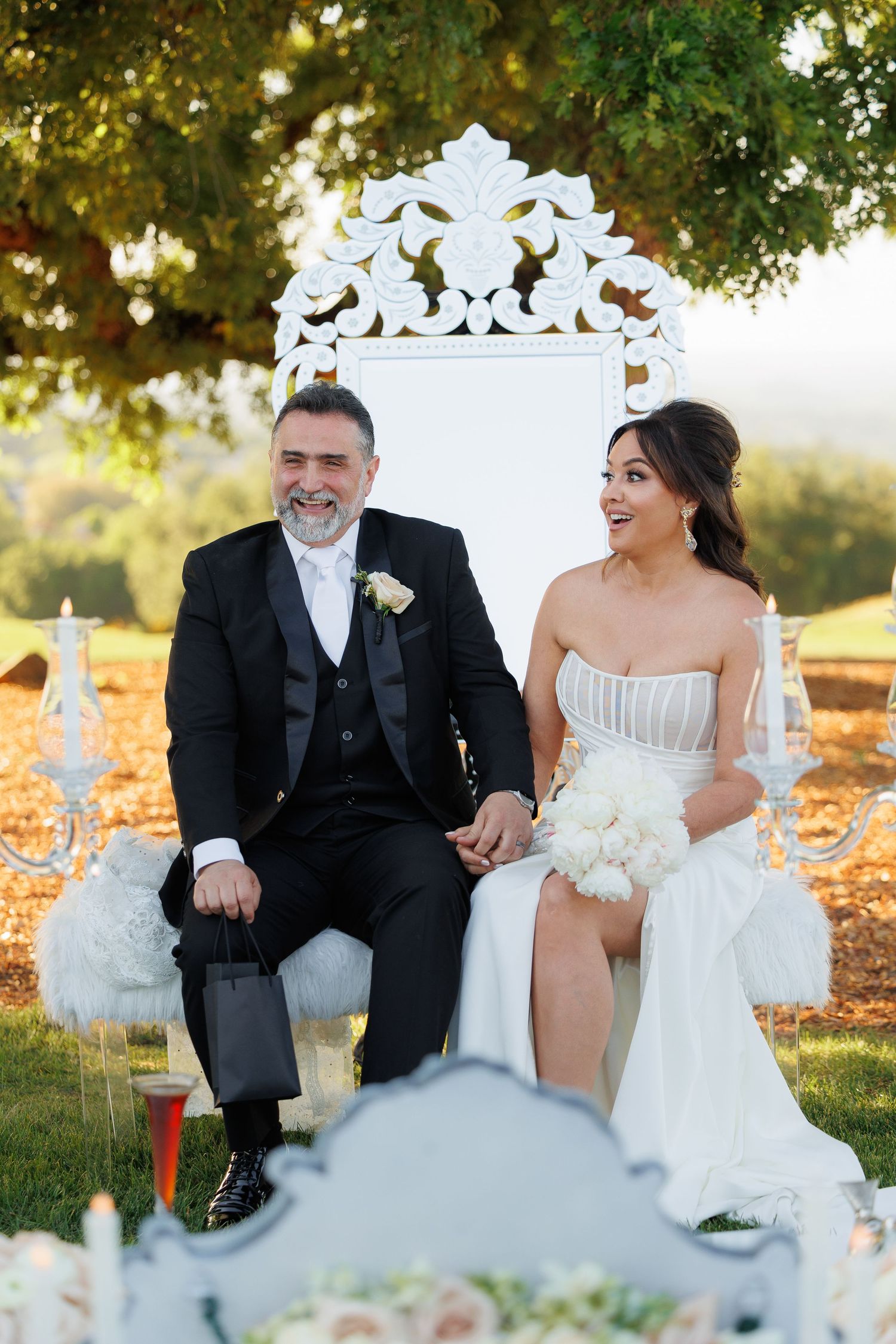 Wedding couple sits together outdoors on white furniture surrounded by autumn leaves at sunset.