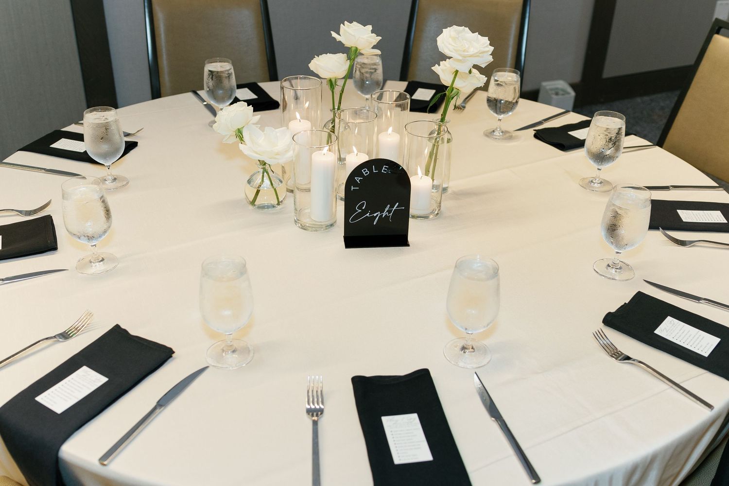 Elegant round dining table set with white linens, black napkins, and delicate white floral centerpieces for a formal event.