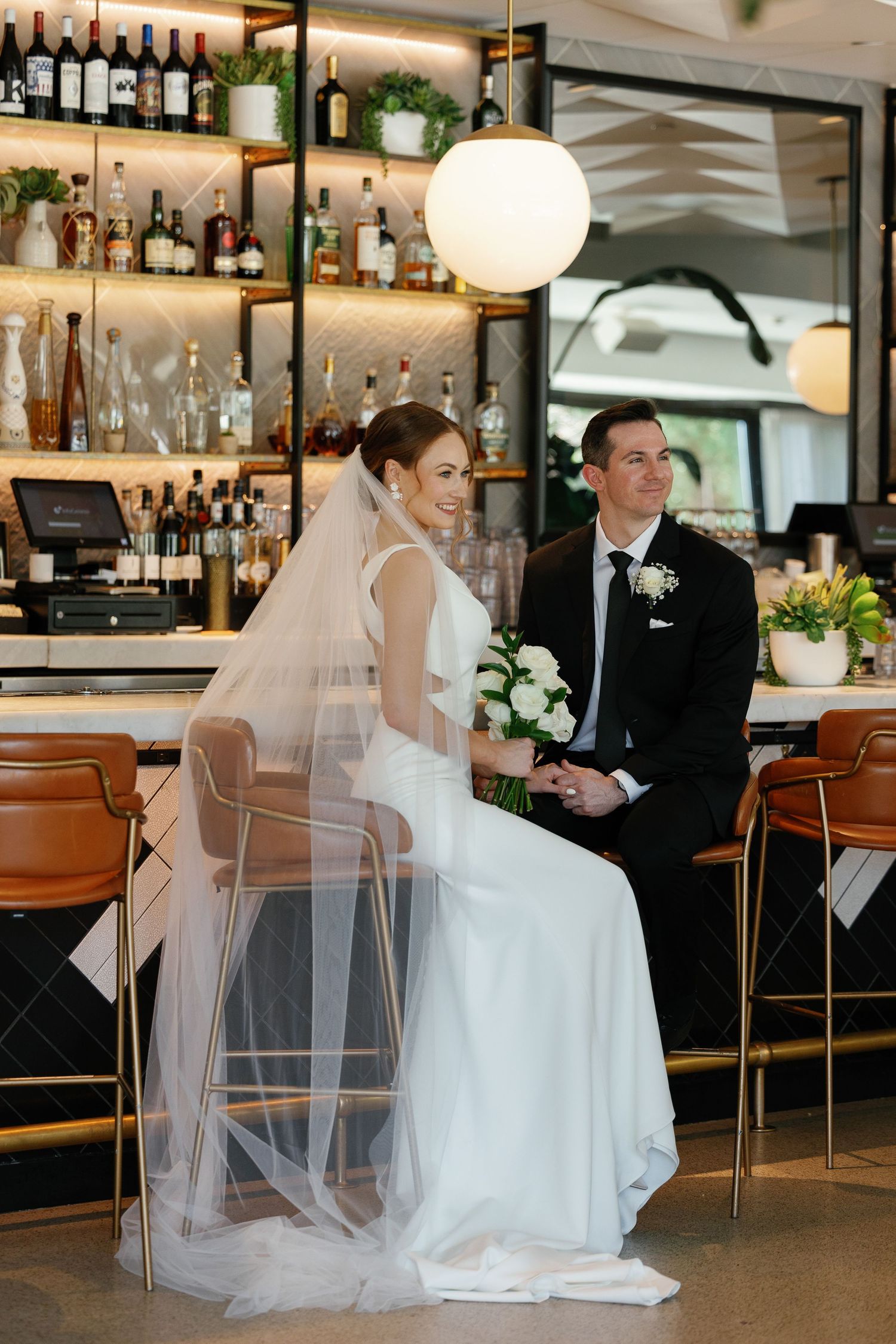 A wedding couple sits at an elegant bar counter in a restaurant with modern decor and lighting fixtures.