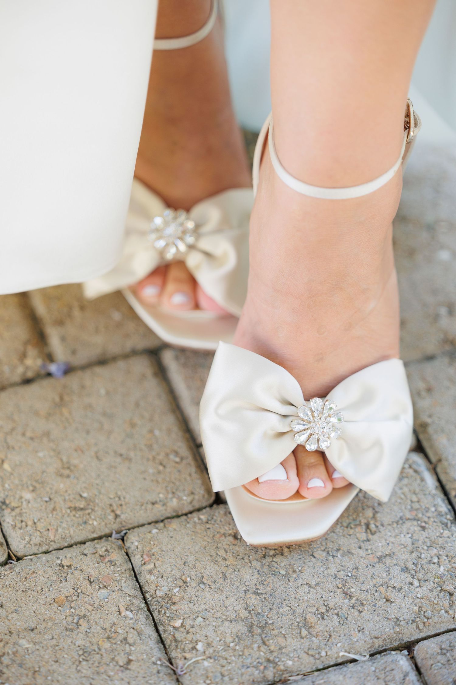 White satin bridal shoes with decorative crystal bow details photographed on stone pavement.