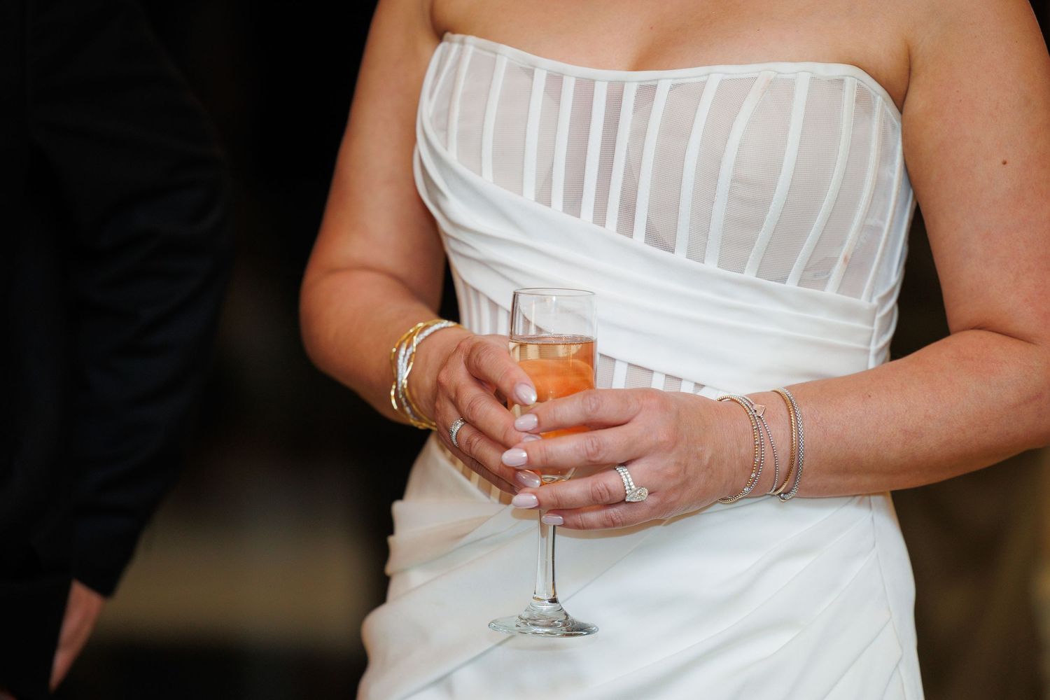 Close-up view of hands holding a champagne glass while wearing an elegant white strapless wedding dress with striped details.