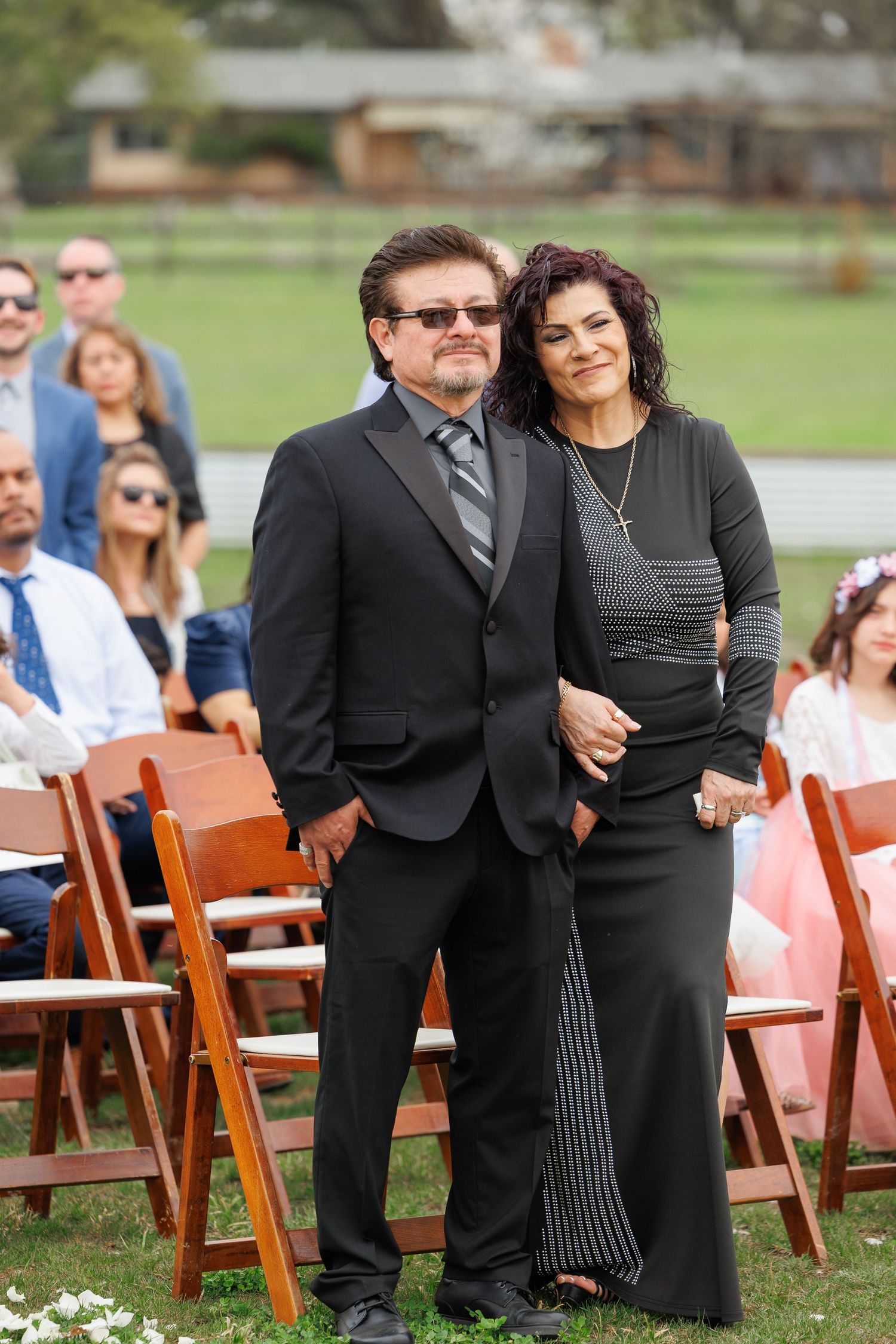 A couple in formal black attire stands together at an outdoor event with wooden chairs and scattered flower petals.